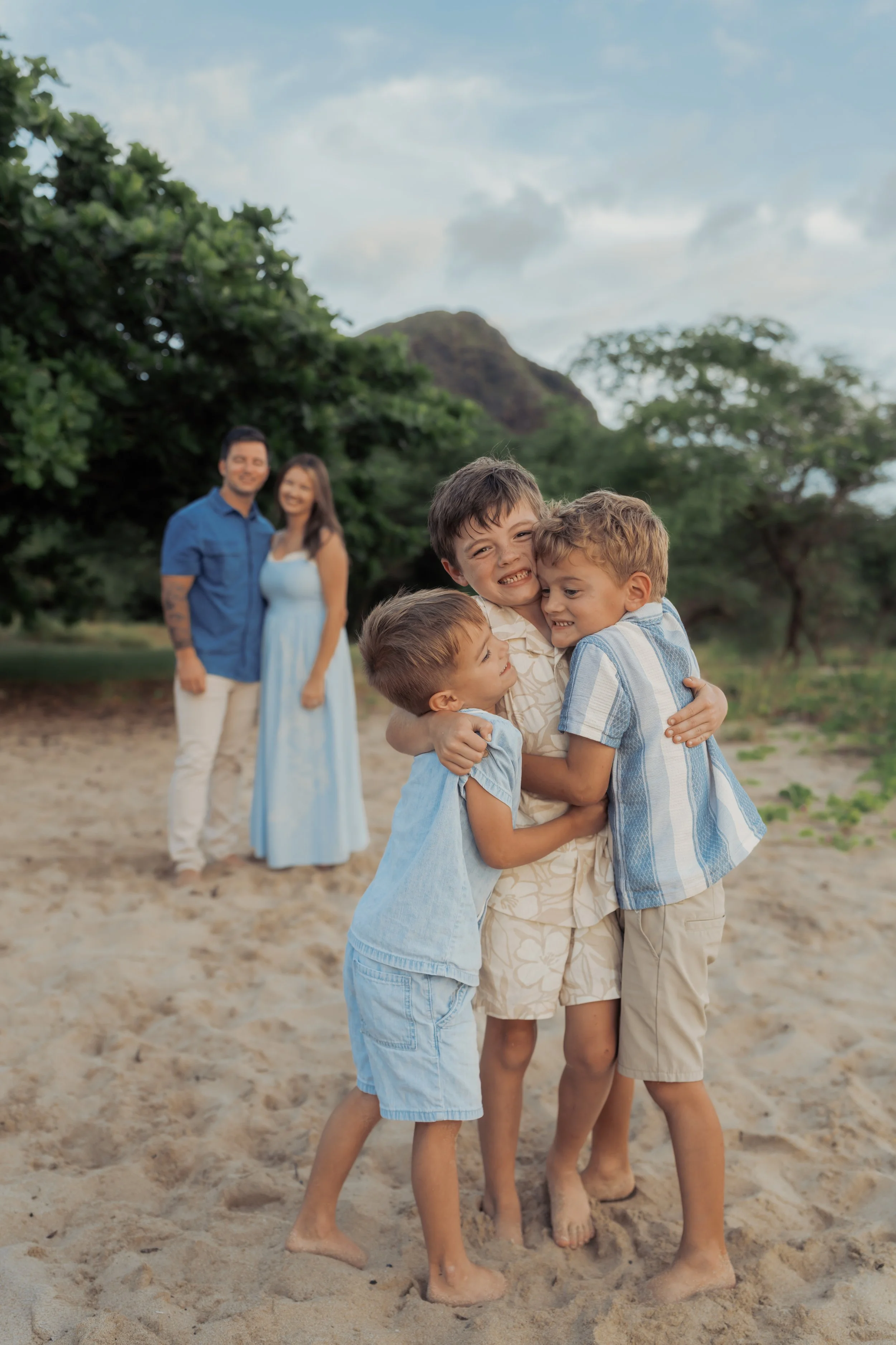 Brothers hug in the foreground on a Hawaiian beach with parents looking lovingly at them