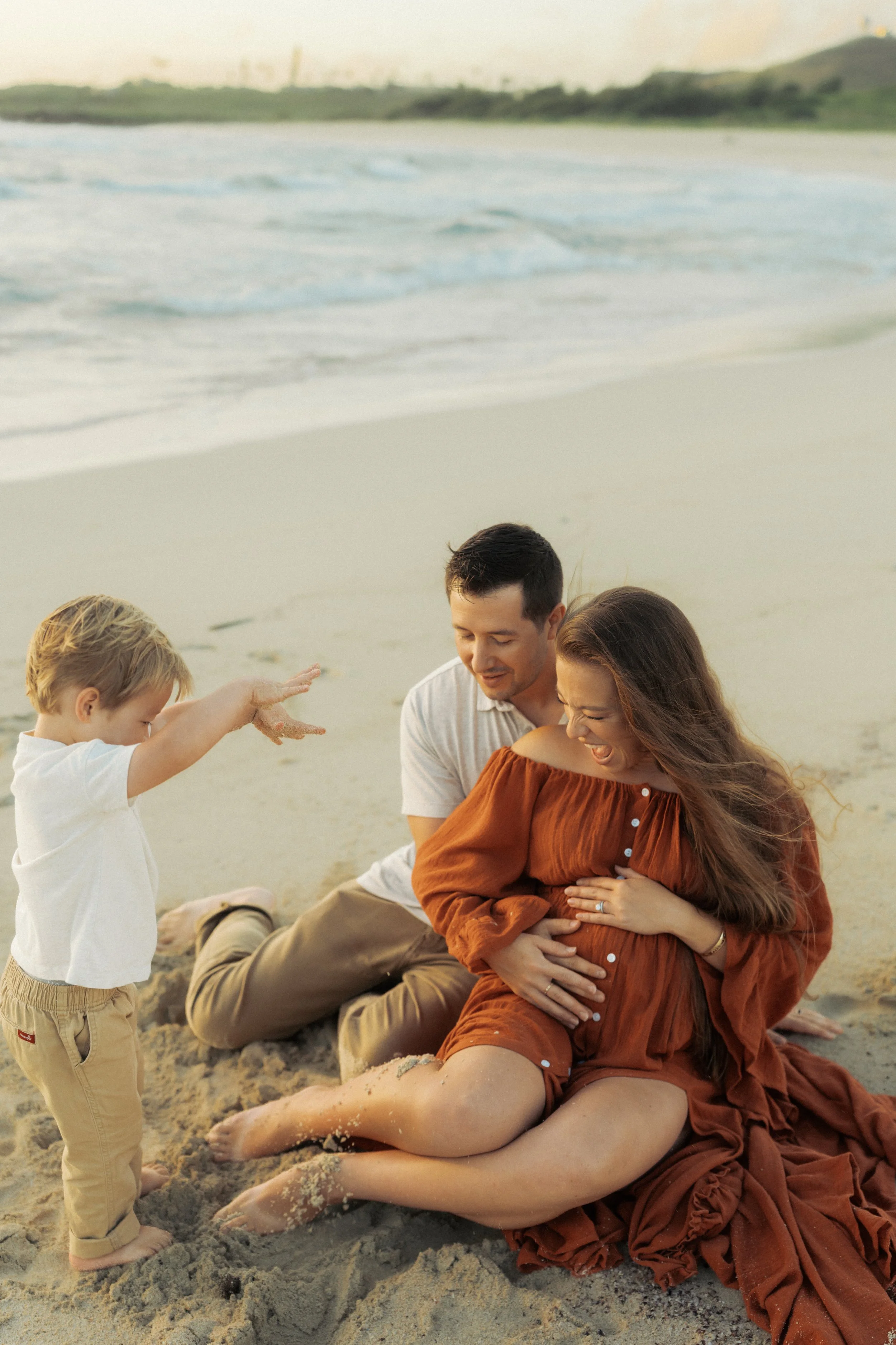 Family maternity session on Oahu beach with toddler throwing sand and playing with parents