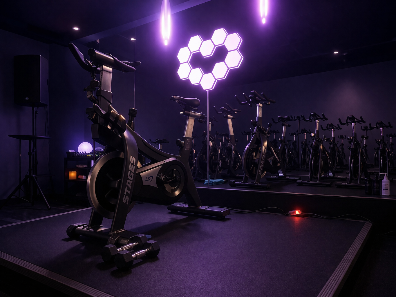Dark indoor gym with stationary bikes in the foreground and a dance or aerobic area with drums and equipment in the background. Colorful lights are projected on the ceiling.