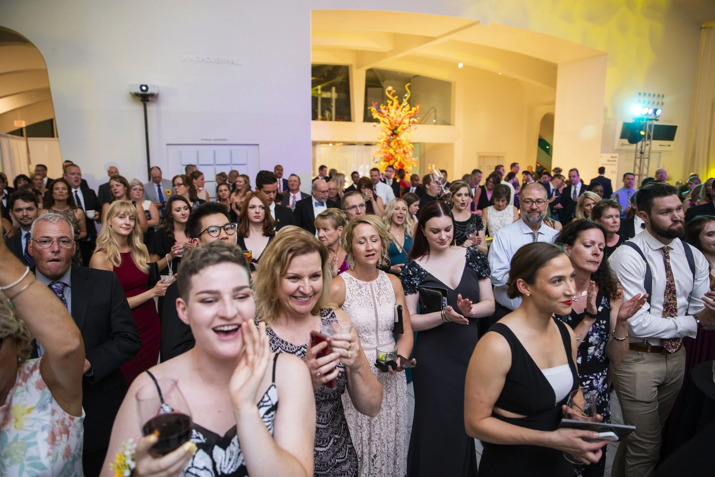 A large group of people dressed in formal attire attending an indoor social event or party, with some smiling and holding drinks.