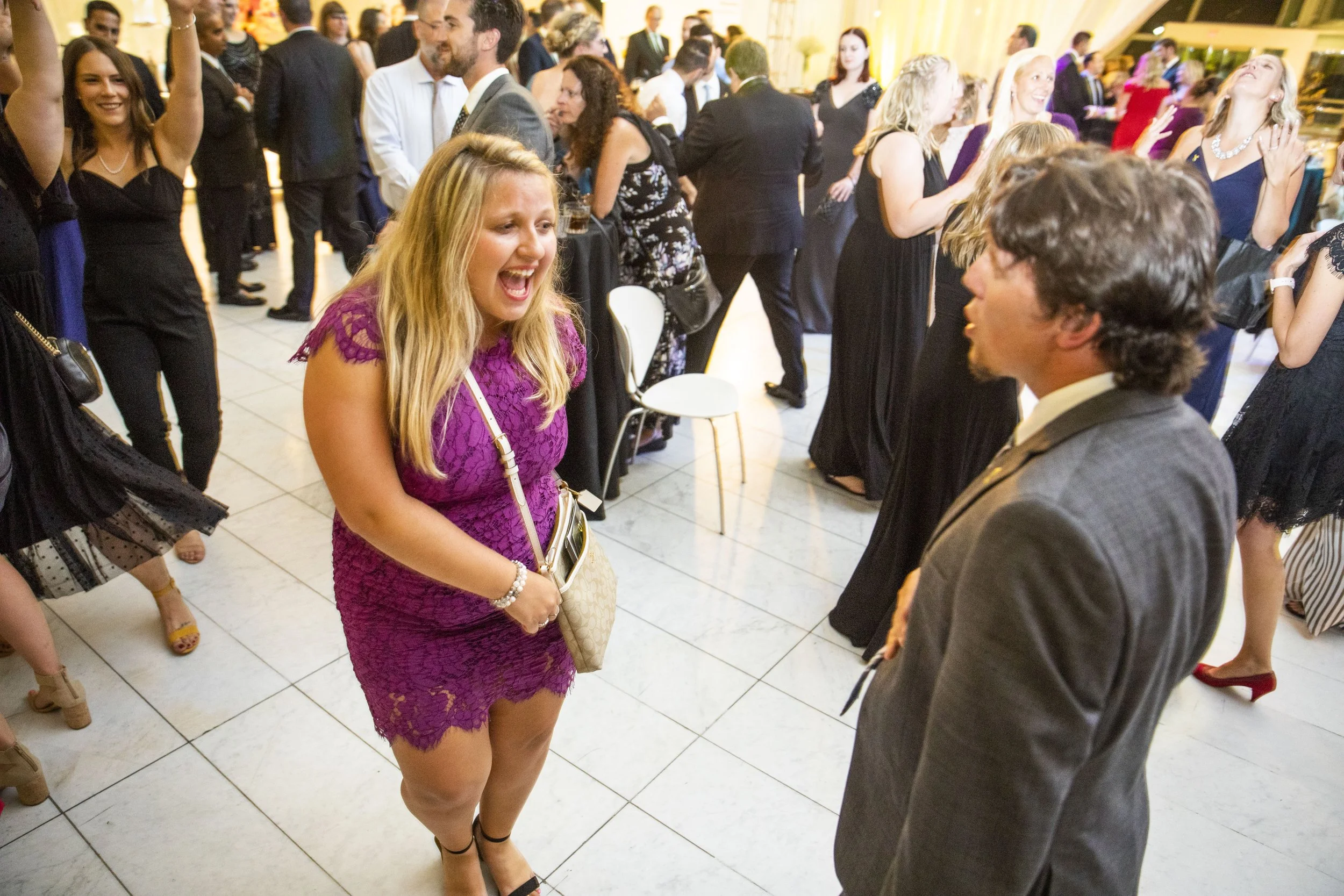 A woman in a purple lace dress is laughing and talking with a man in a gray suit at a formal event, with other guests dancing and mingling in the background.