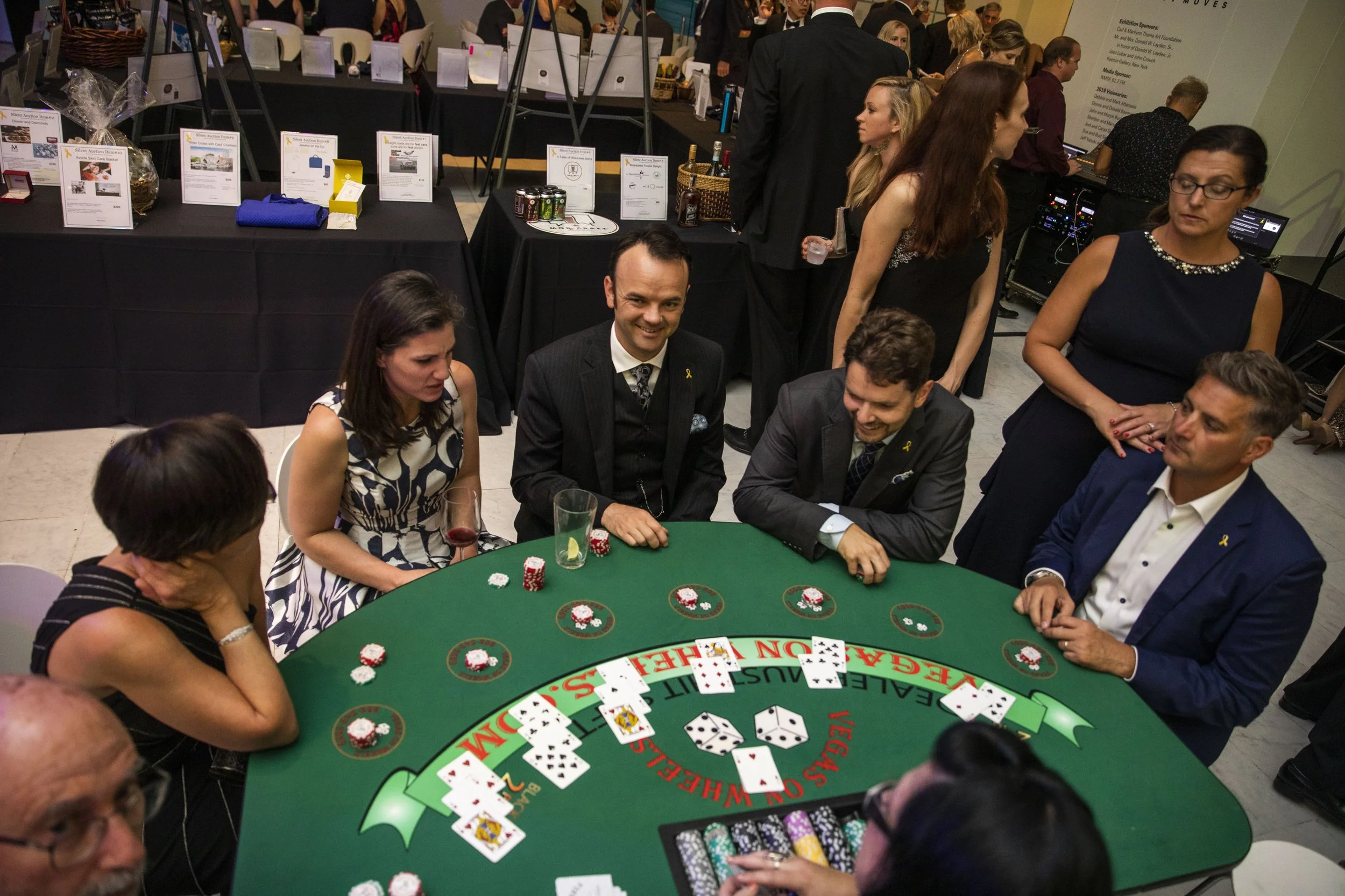 People playing blackjack at a casino table during an event, with chips and cards on the table and others in the background.