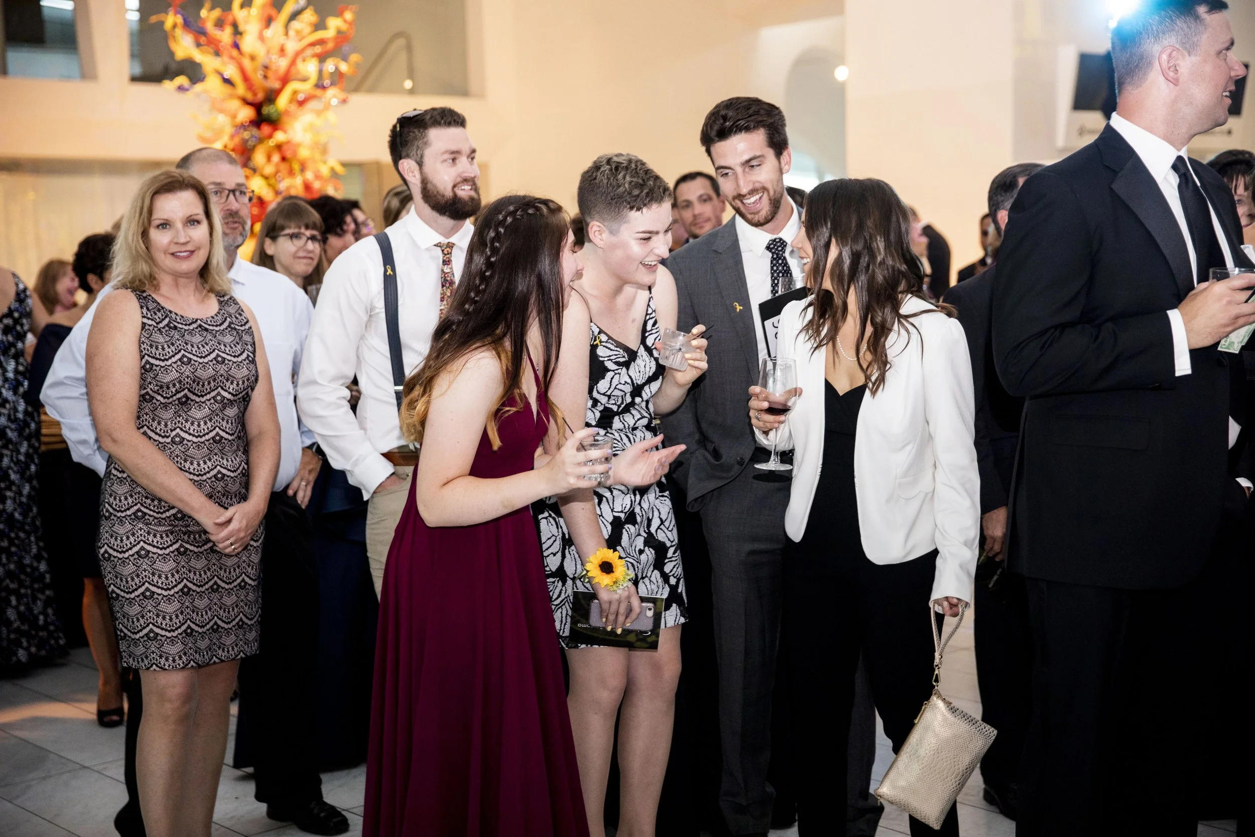 Group of people dressed in formal attire socializing at a reception with a bright floral centerpiece in the background.