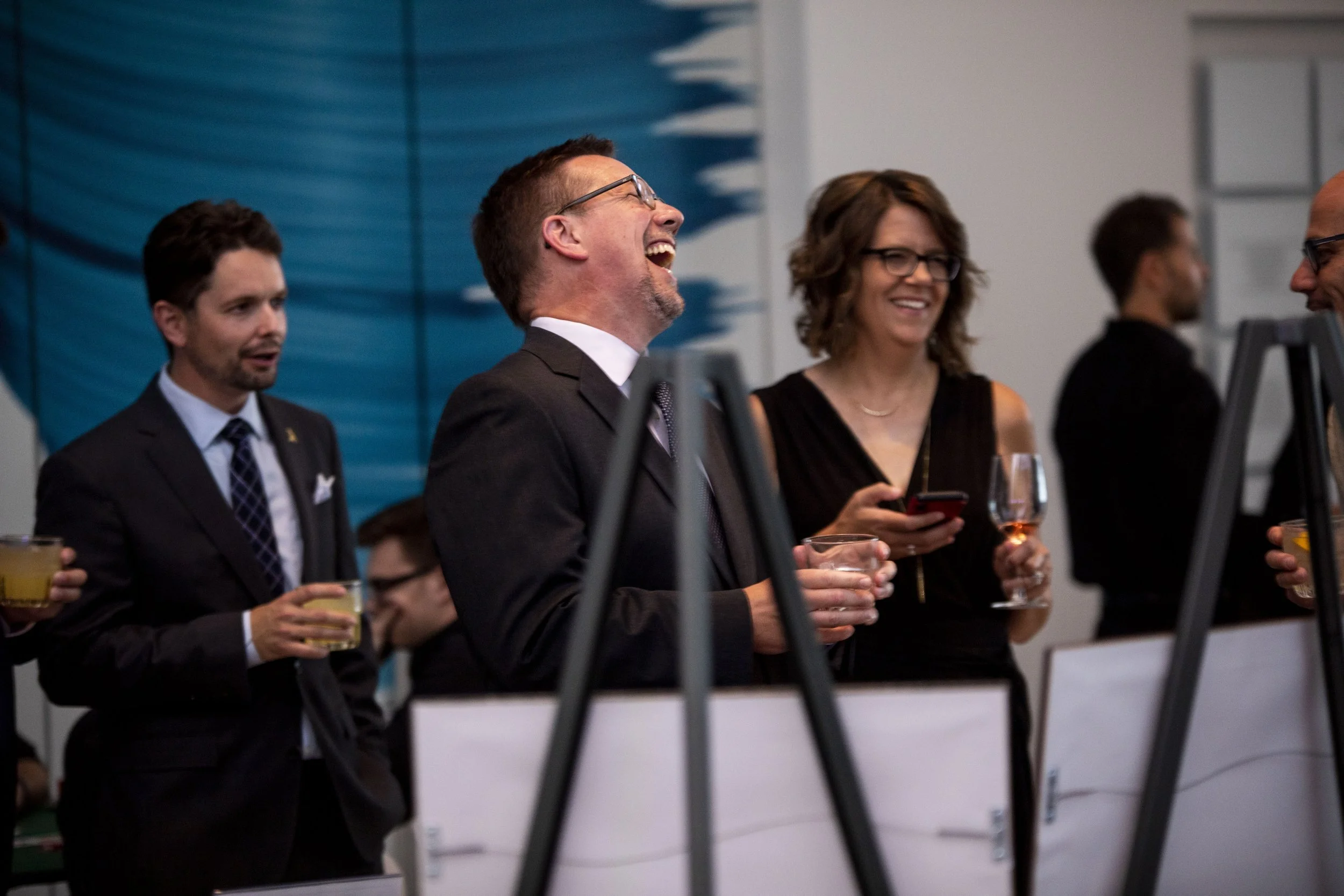 A group of professionally dressed people at a social event, holding drinks and engaging in conversation, with some smiling and laughing.