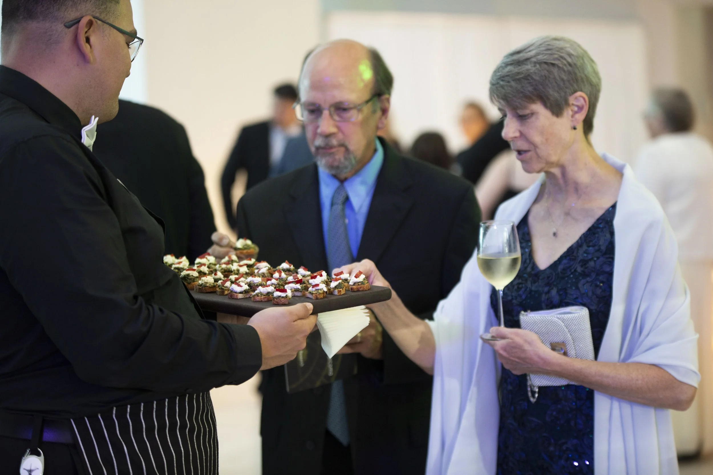 A woman with short gray hair holding a glass of white wine and a purse, reaching for a tray of hors d'oeuvres at a social event. A man with glasses and a woman in a dark blue dress with a white shawl are also looking at the tray. There are other peop