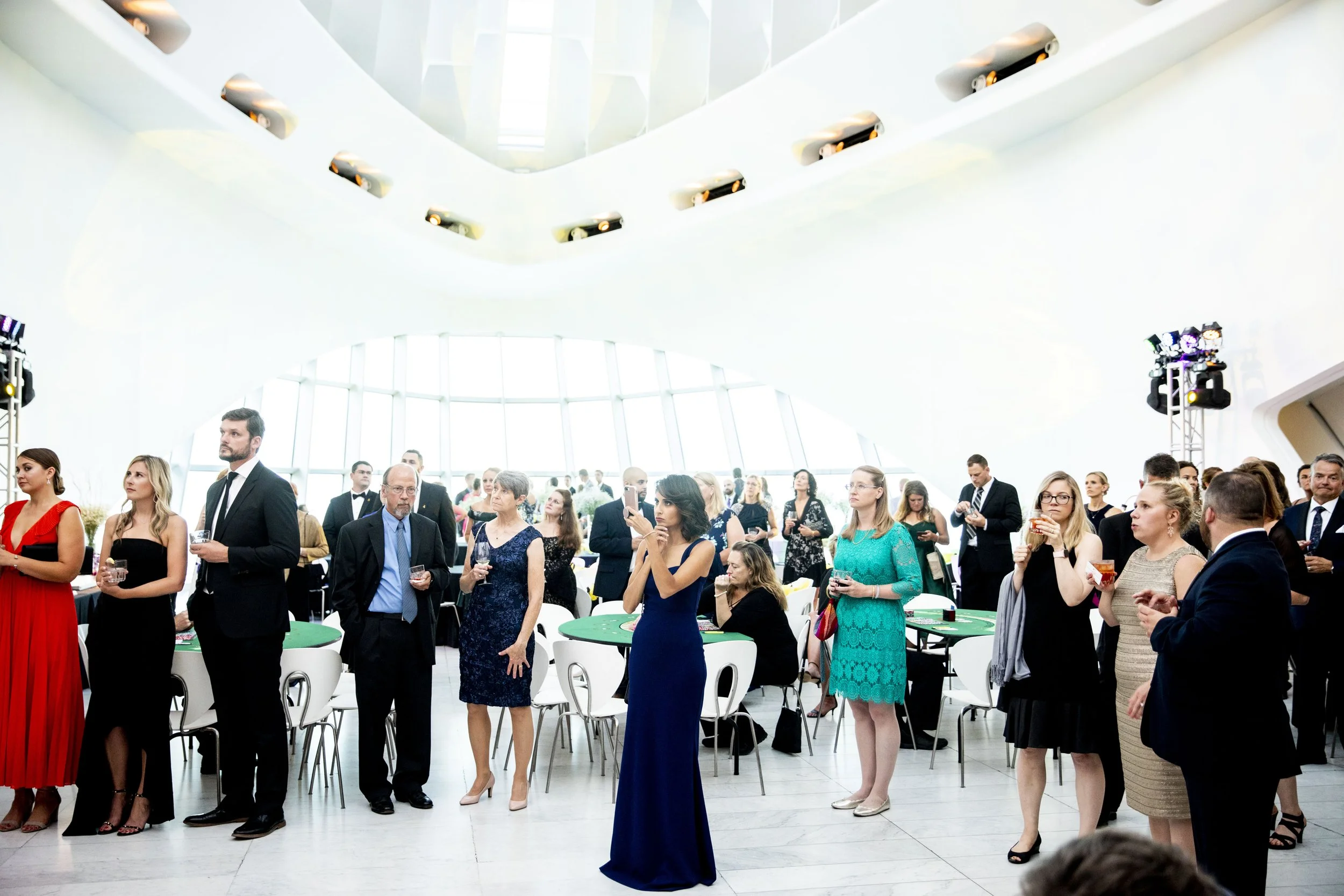 Group of people dressed in formal attire at a social event or reception inside a modern, well-lit building with large windows and white architecture.