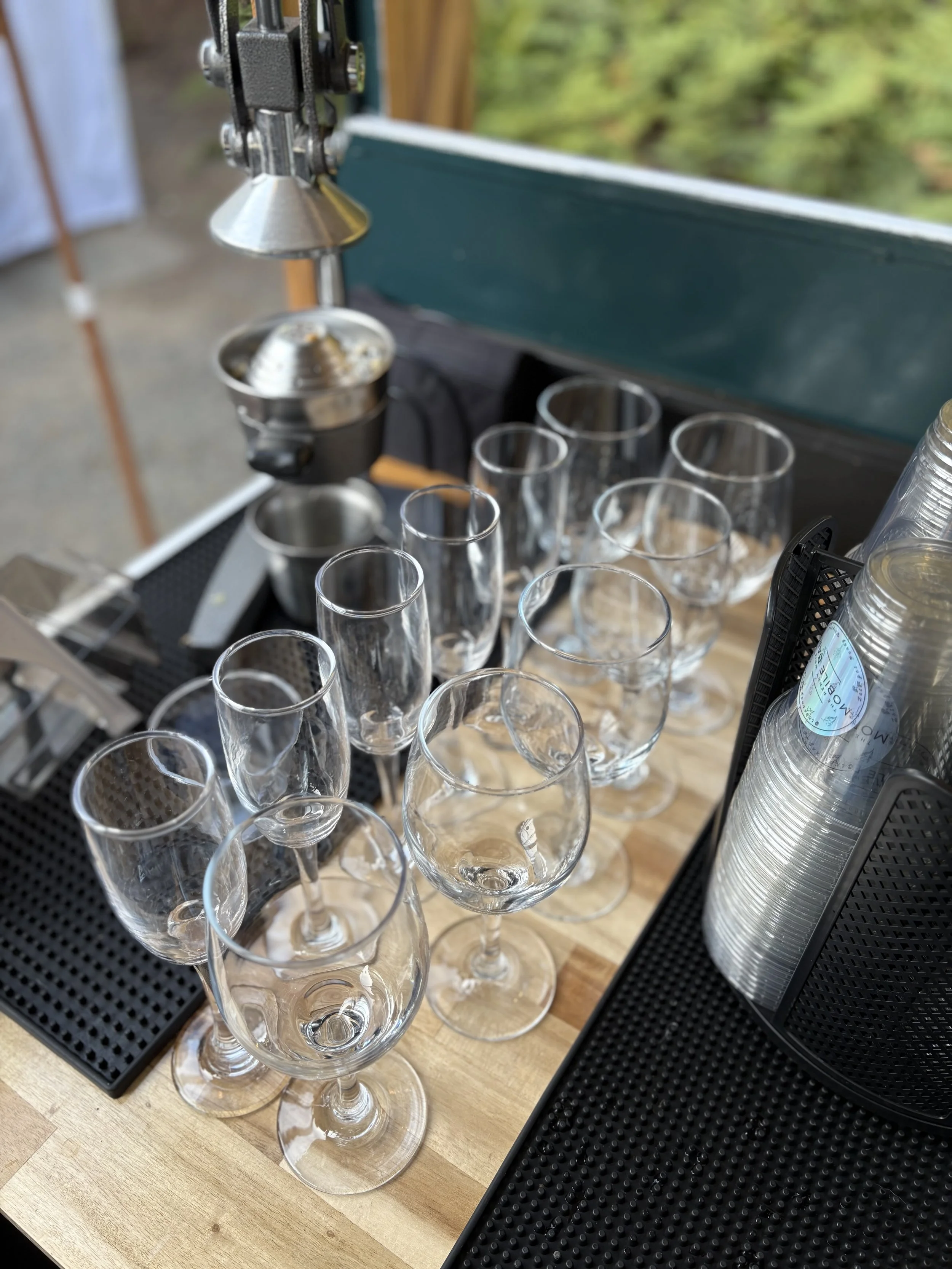 Multiple empty wine glasses on a wooden bar counter next to a stack of disposable cups and a black organizing tray.