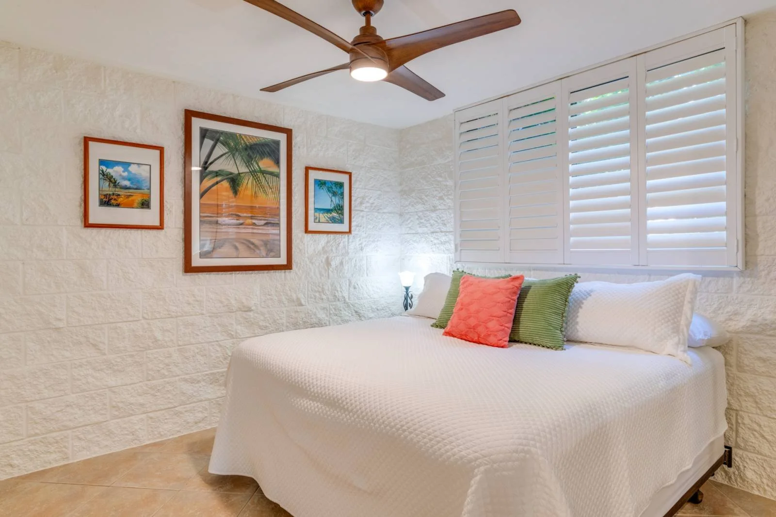 A bedroom with a white bedspread, multiple pillows in shades of orange, green, and white, and a window with white shutters. There are three framed pictures of tropical beach scenes on the wall, a ceiling fan, and a small bedside lamp.