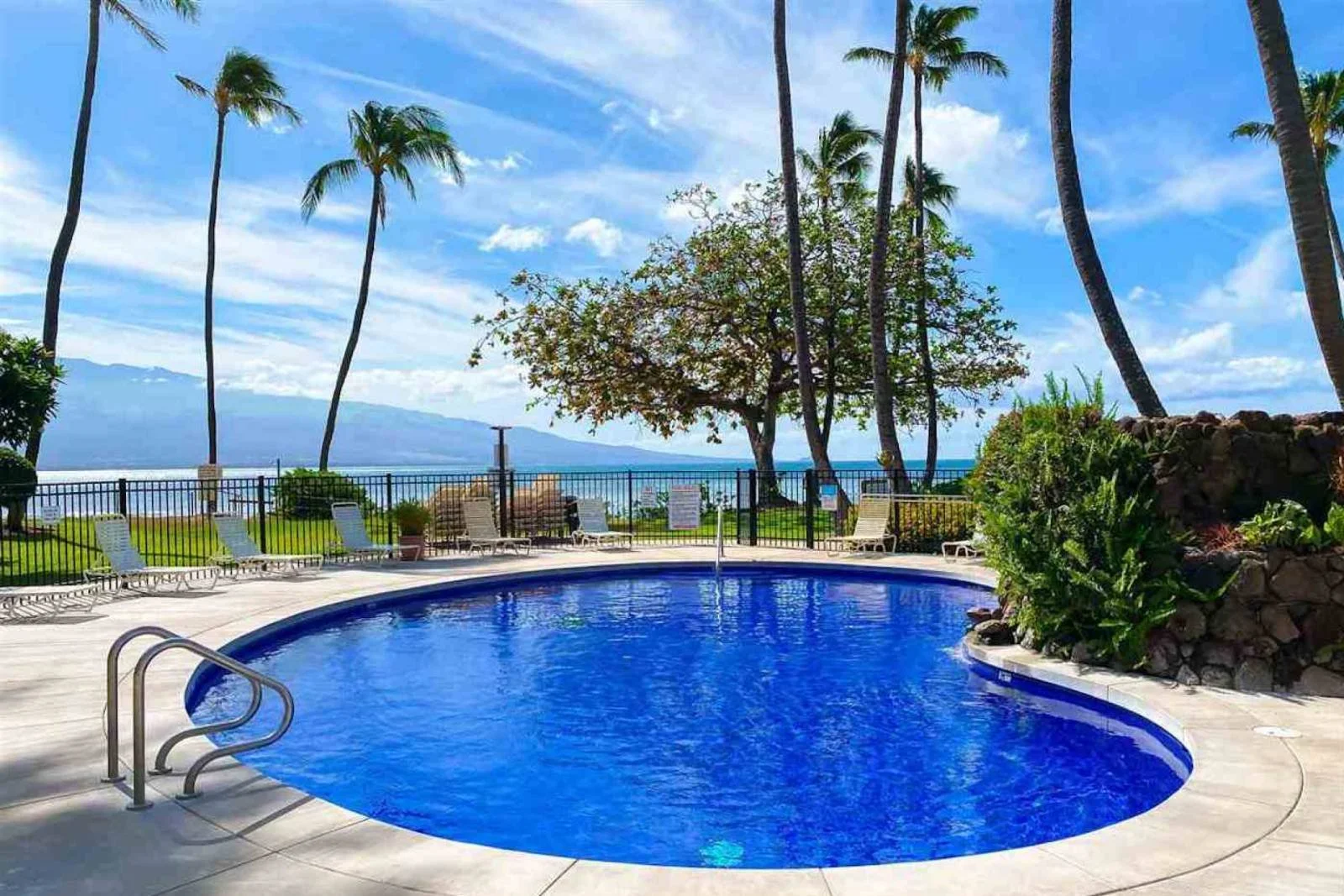 A tropical outdoor swimming pool area with blue water, surrounded by lounge chairs and lush green plants, overlooking the ocean and mountains in the distance, under a partly cloudy sky with palm trees.
