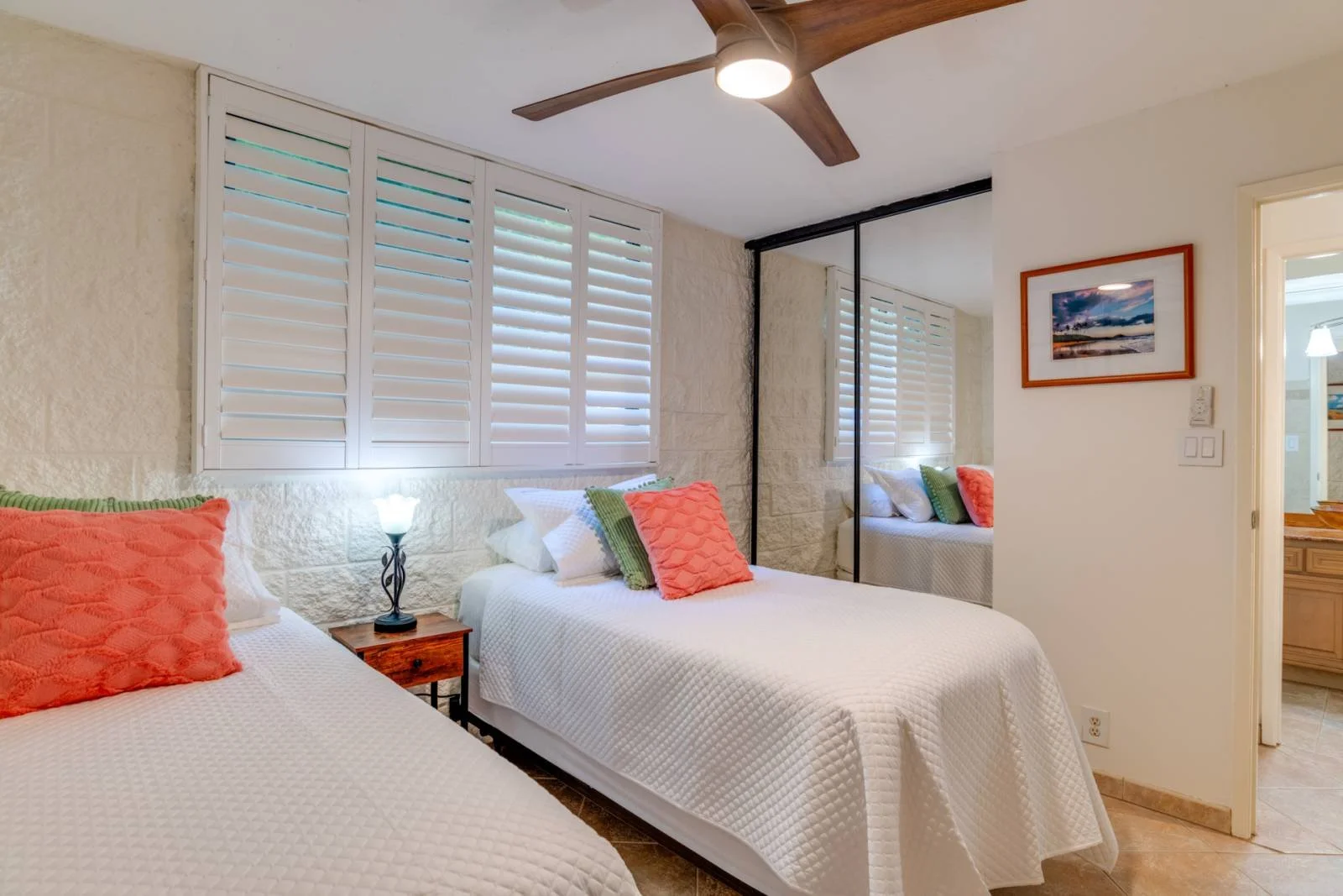 A bedroom with two beds covered in white quilts, each with pink and green pillows. There is a small wooden nightstand with a lamp between the beds. The room has a stone wall with white shutters covering the windows, and a mirrored closet reflective o