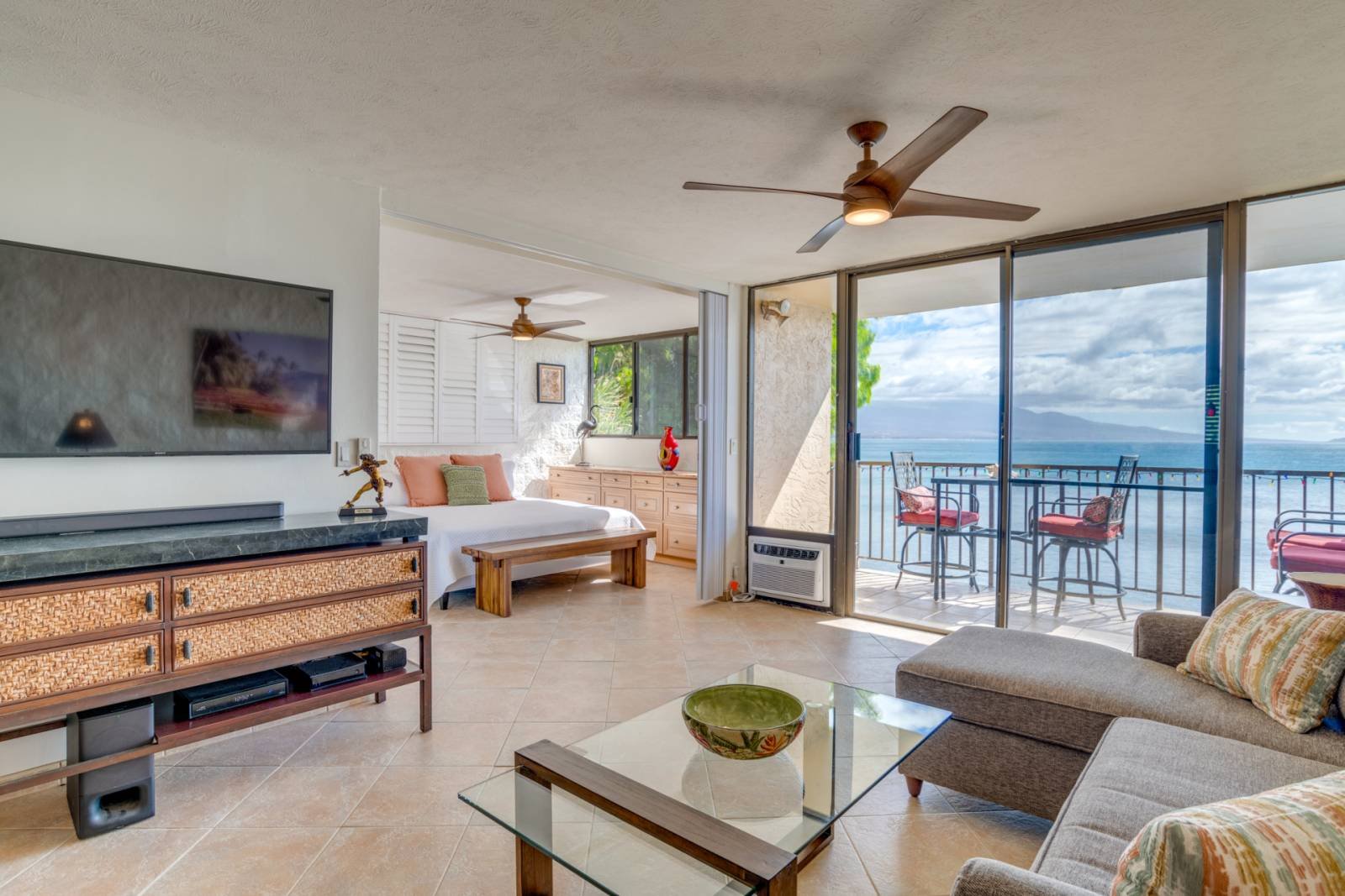 Living room with sliding glass door leading to balcony with ocean view, featuring a sofa, glass coffee table, wall-mounted TV, ceiling fans, and a bedroom area in the background.
