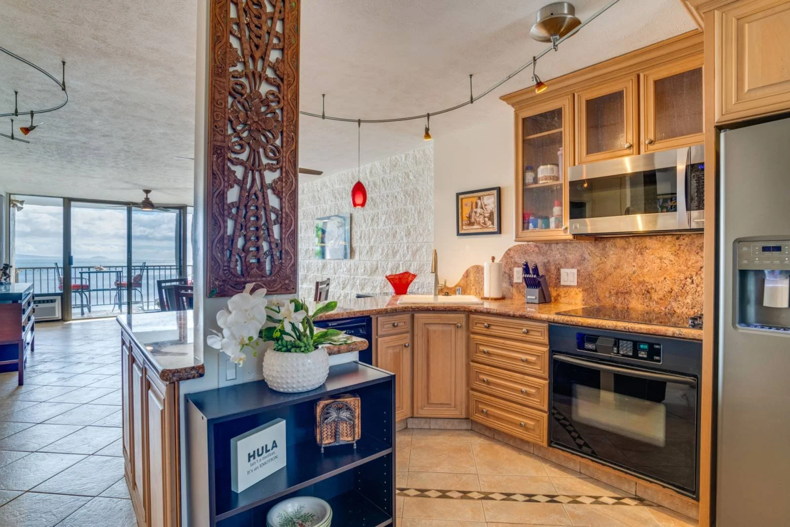 Interior view of a kitchen with wooden cabinets, a granite counter, a metal microwave, and a refrigerator. Visible living room area with large windows and a balcony in the background.