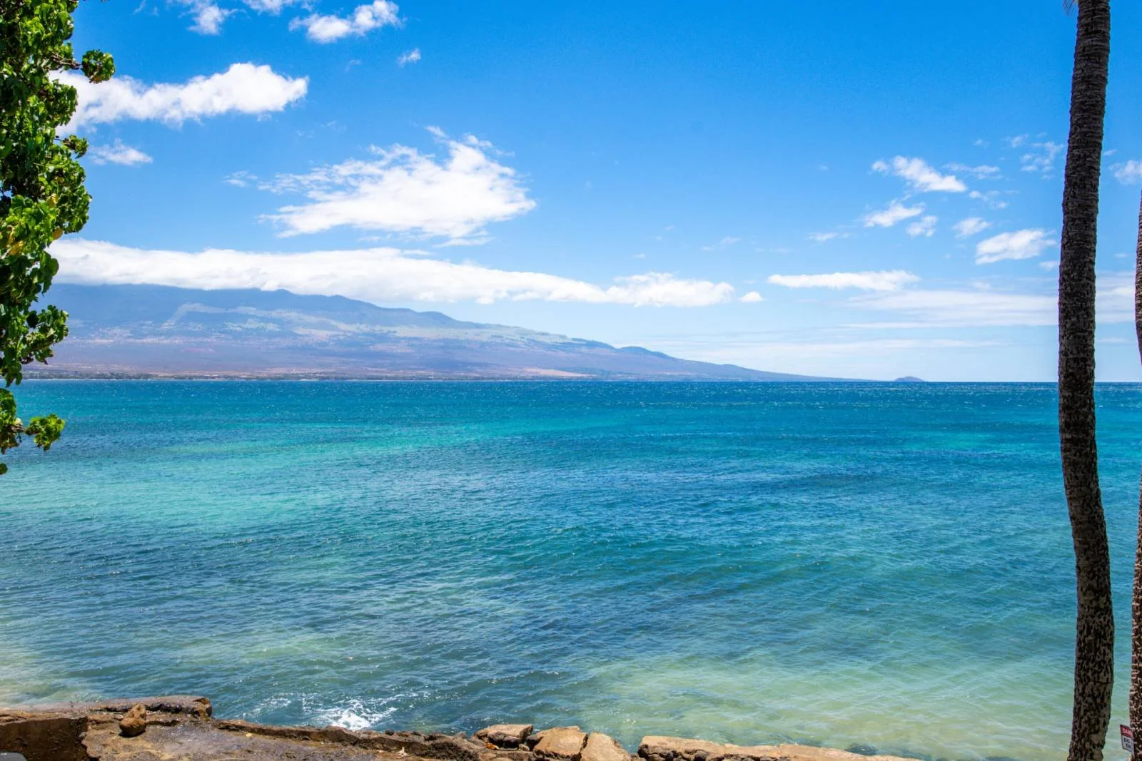 Tropical beach with clear blue water, palm trees, and distant mountains under a partly cloudy sky.