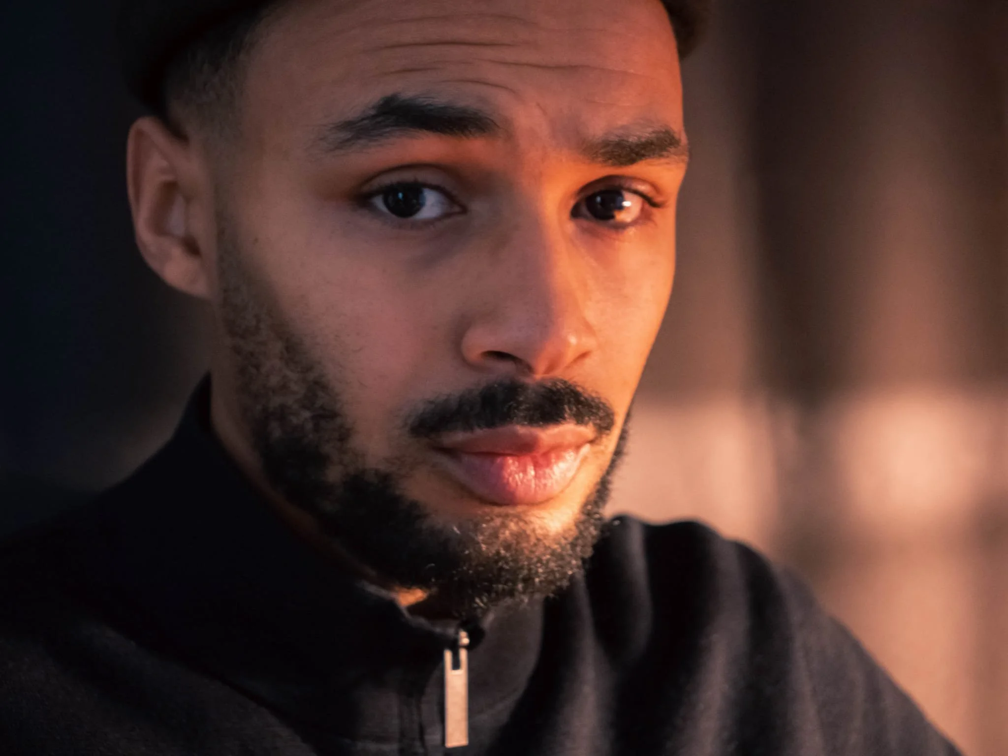 A close-up portrait of a man with short dark hair, brown eyes, a beard, and mustache, wearing a black zip-up jacket, looking directly at the camera with a neutral expression, illuminated by soft warm lighting.