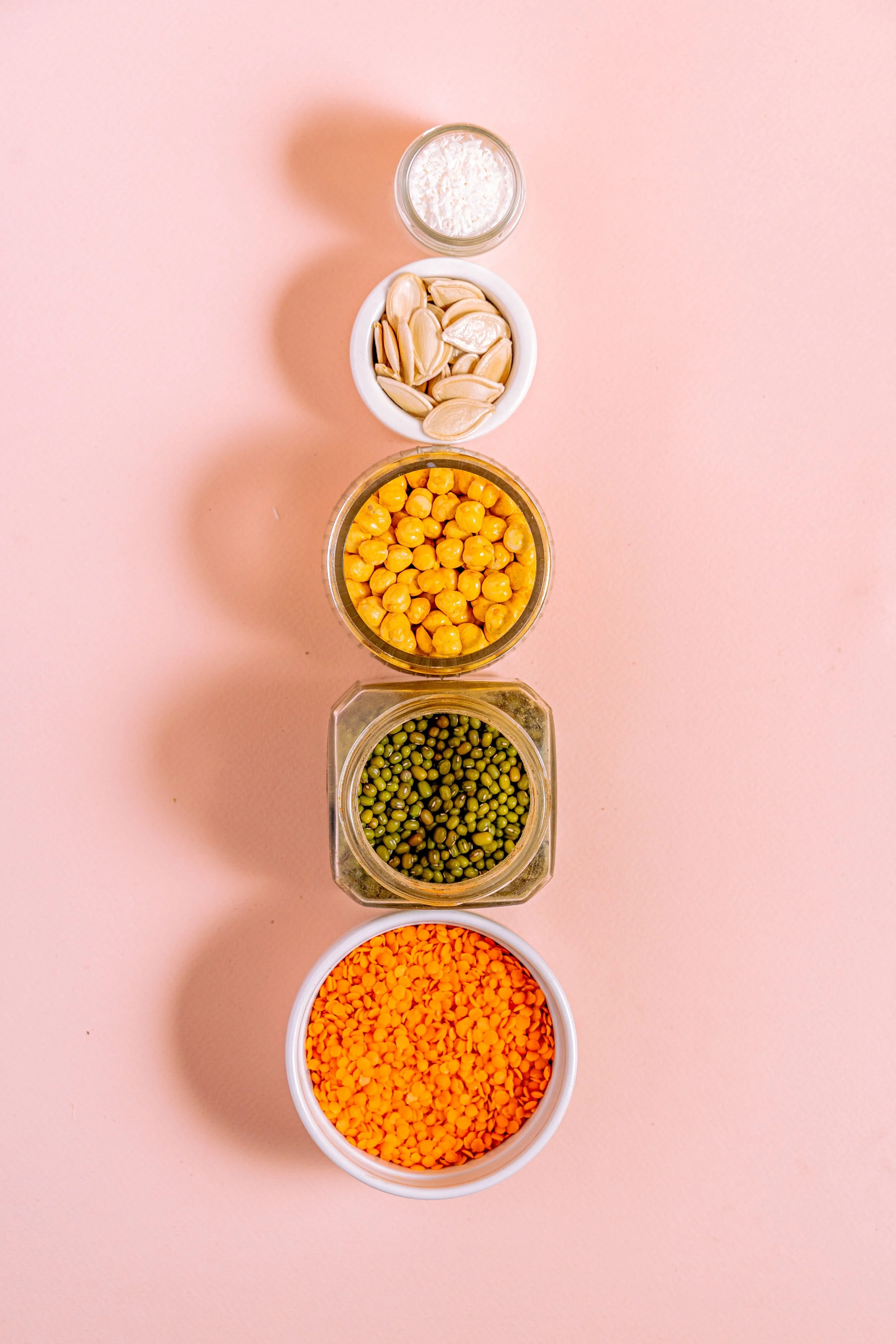 Bowl and jars of dried pantry staples arranged in a line. Top to bottom pumpkin seeds, chickpeas, mung beans, and red lentils.