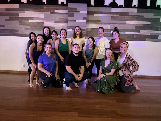 A group of 14 individuals posing together in a room with wooden flooring and a patterned wall. They appear to be a diverse mix of men and women smiling at the camera, some standing and some kneeling.