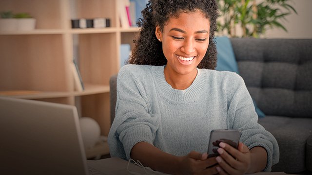 Smiling woman using smartphone at home office with laptop and bookshelf in background