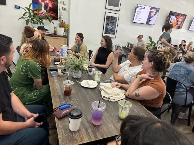 A group of people sitting around a long wooden table in a cafe or coffee shop. They are engaged in conversation and drinks are placed on the table. The setting is relaxed and informal, with plants and posters decorating the room.