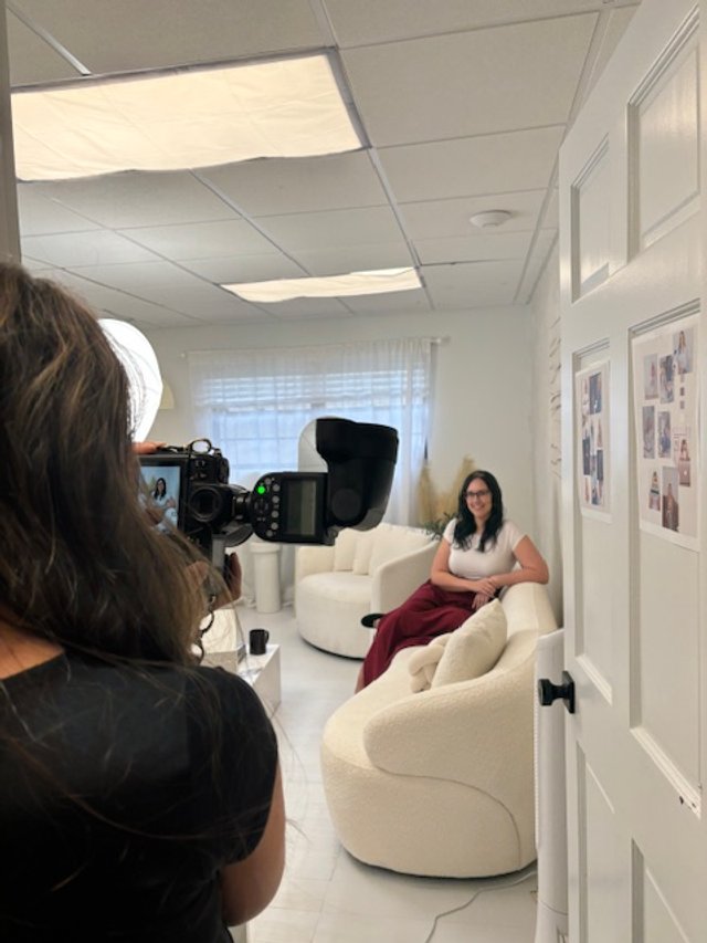 Photo shoot setup with a woman sitting on a white couch, being photographed in a well-lit room.