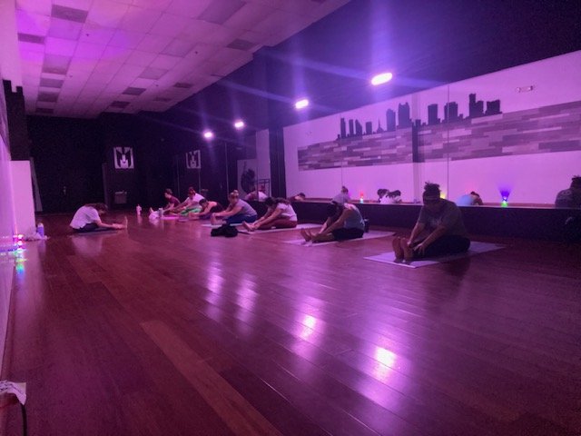 People doing yoga stretches in a dimly lit room with wooden flooring, purple lighting, and a mirrored wall featuring a city skyline mural.