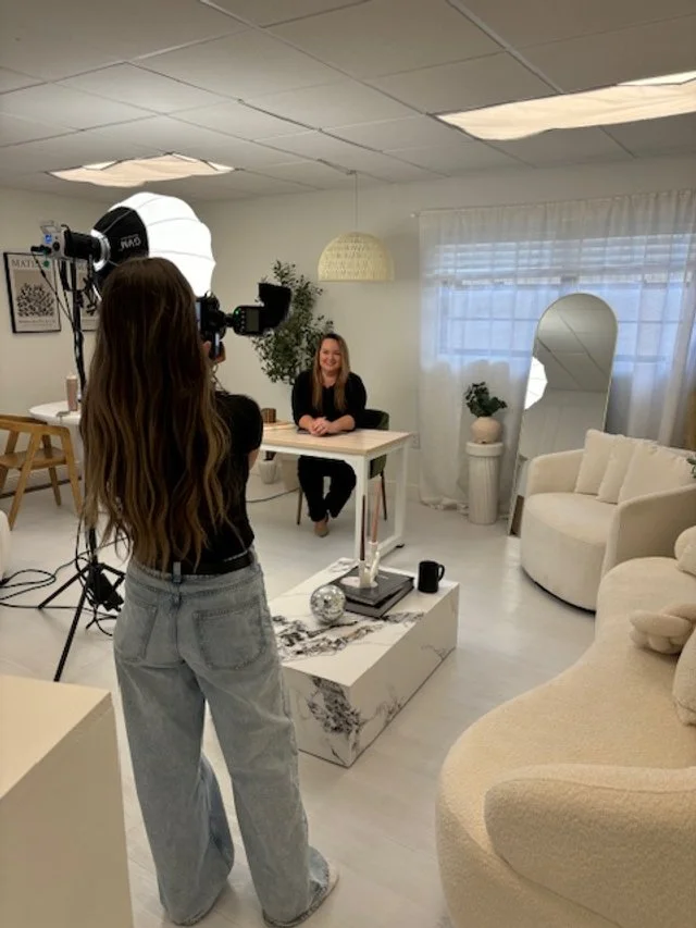 A photographer taking a picture of a woman sitting at a desk in a well-lit, modern office space with a cozy seating area and decorative plants.