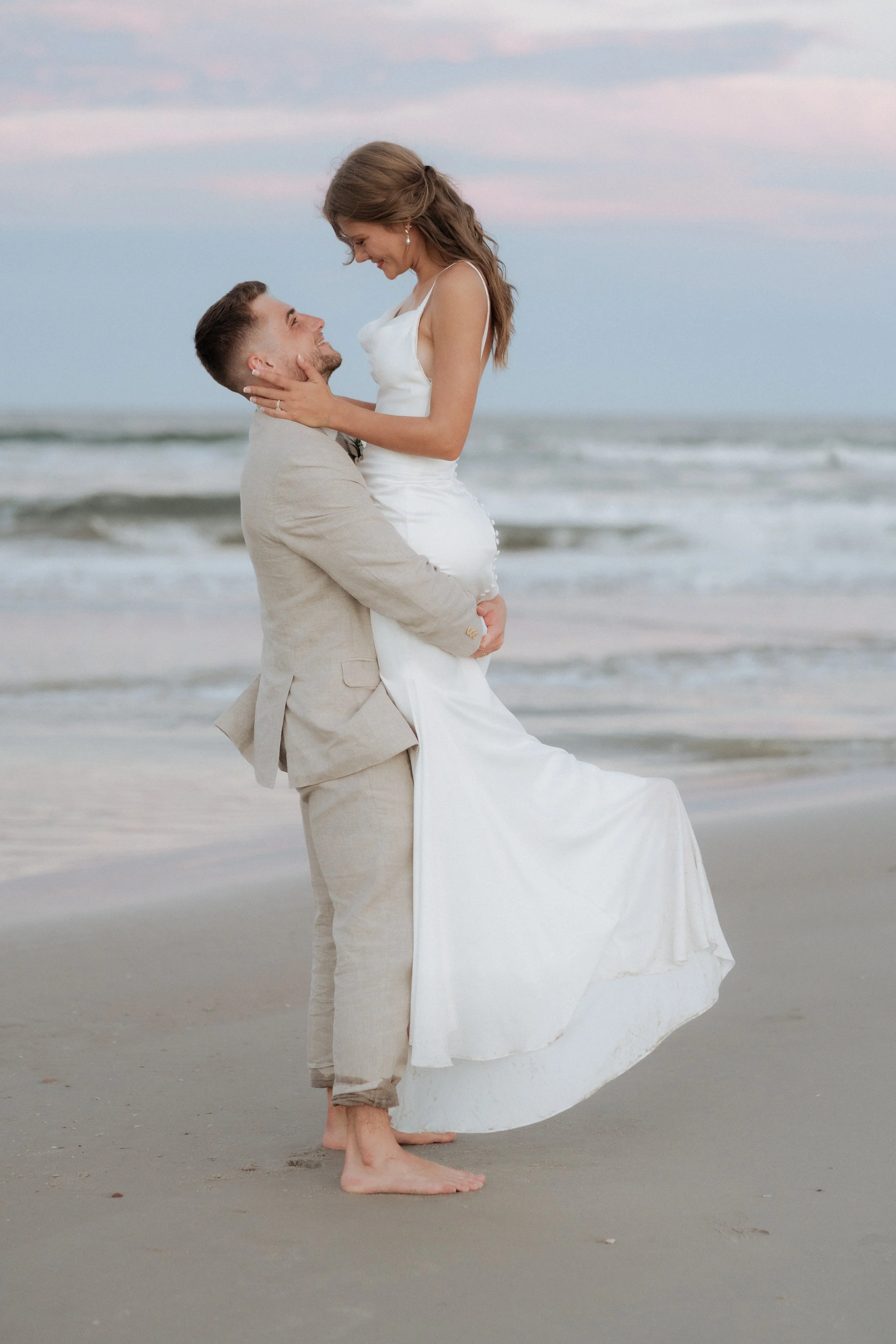 A couple in wedding attire on the beach at sunset, with the man lifting the woman while they smile at each other.