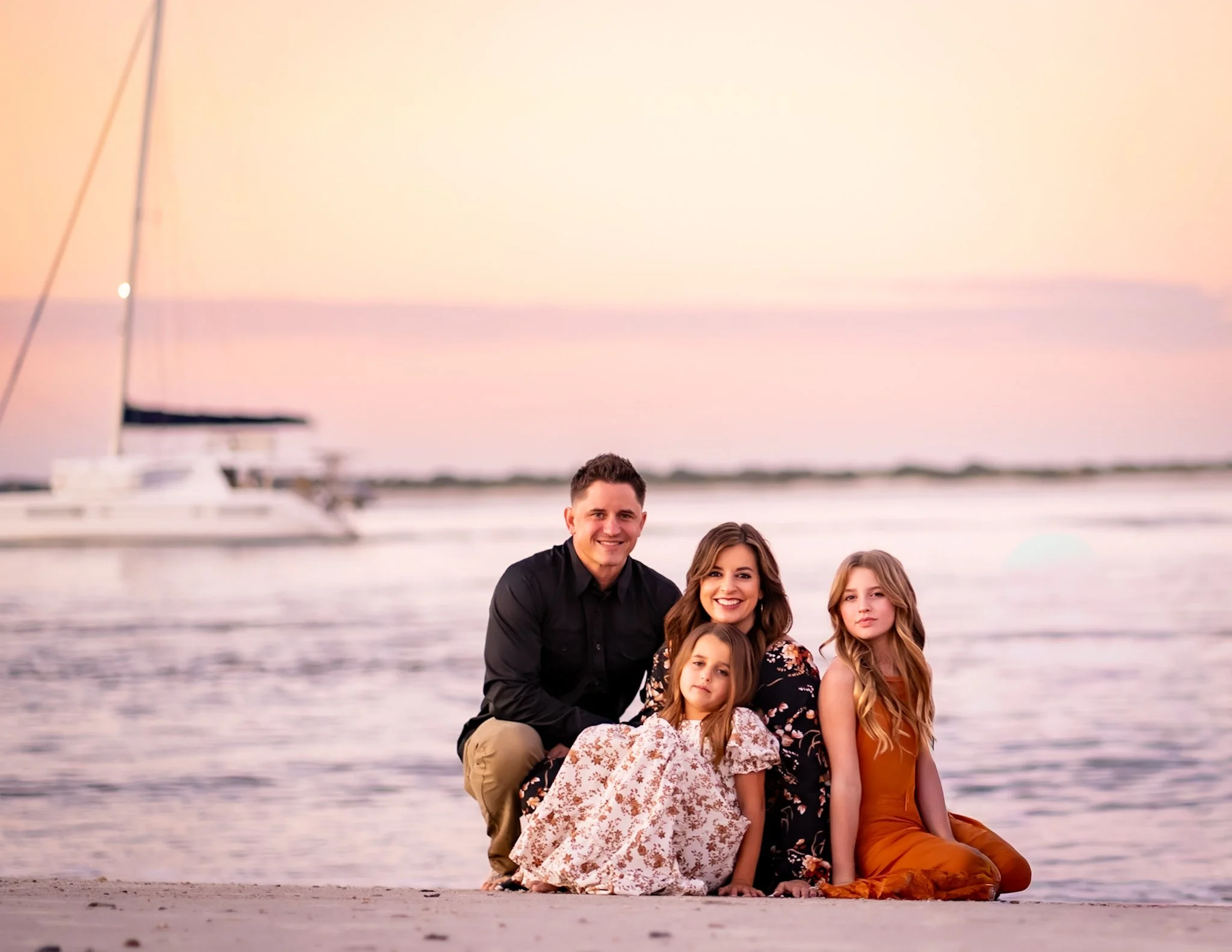 A mom, dad and daughters sit on a beach on Amelia Island Florida for their family pictures to be taken. A sailboat is in the background of the picture to show the beauty of the island.