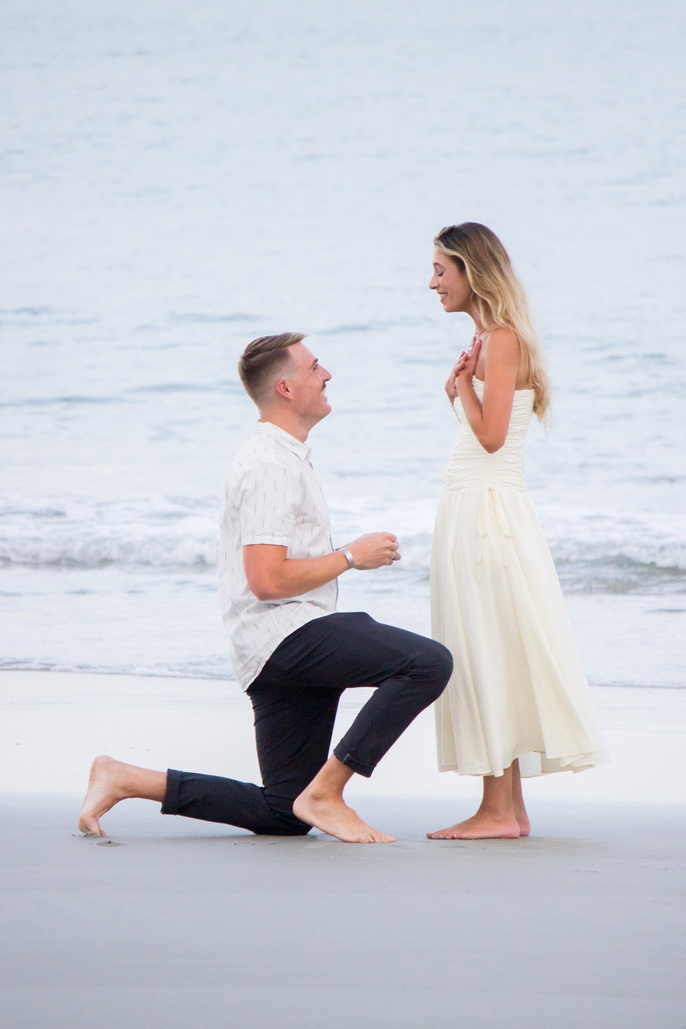 A man surprise proposes to his girlfriend on Amelia Island at The Ritz Carlton Amelia Island. She is surprised and is so excited while she stands on the sand and he is on one knee in front of the blue ocean waves.