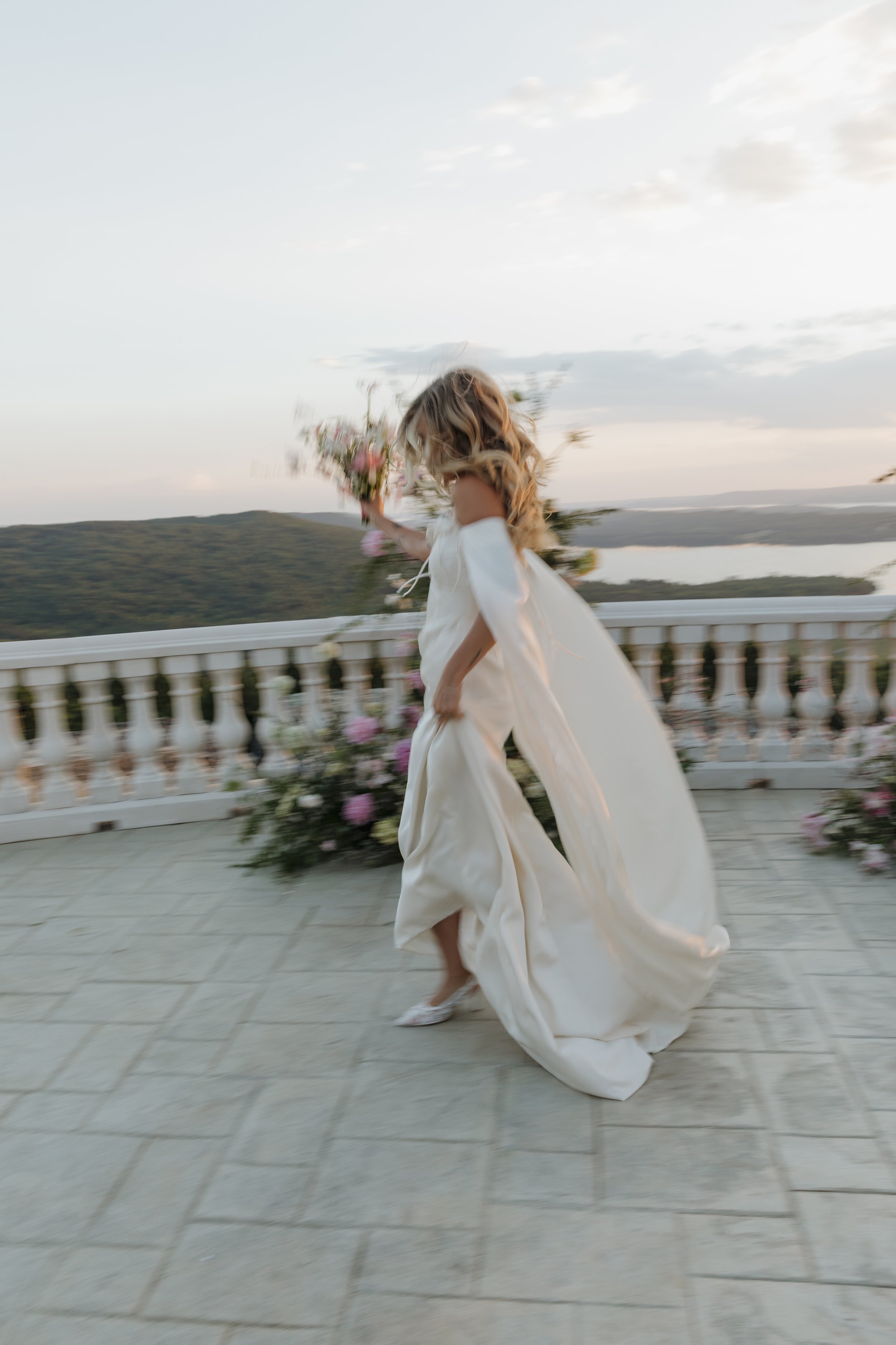 A woman in a flowing cream-colored dress holding a bouquet of flowers, running on a balcony with a scenic view of hills and a lake at sunset.