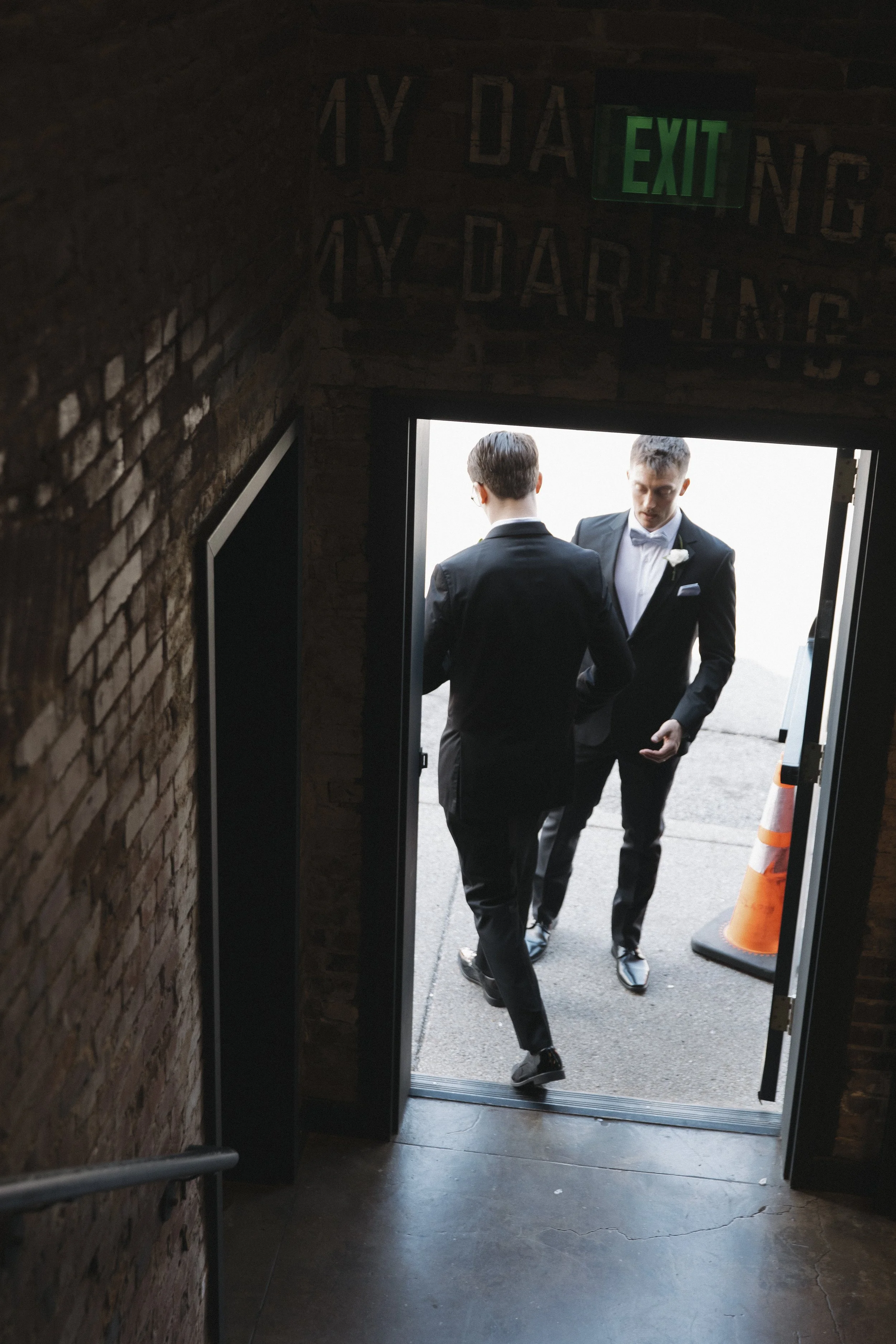 Two young men in tuxedos entering a building through a black doorway, with an orange traffic cone nearby, seen from inside a brick-walled room. An illuminated green EXIT sign is above them.