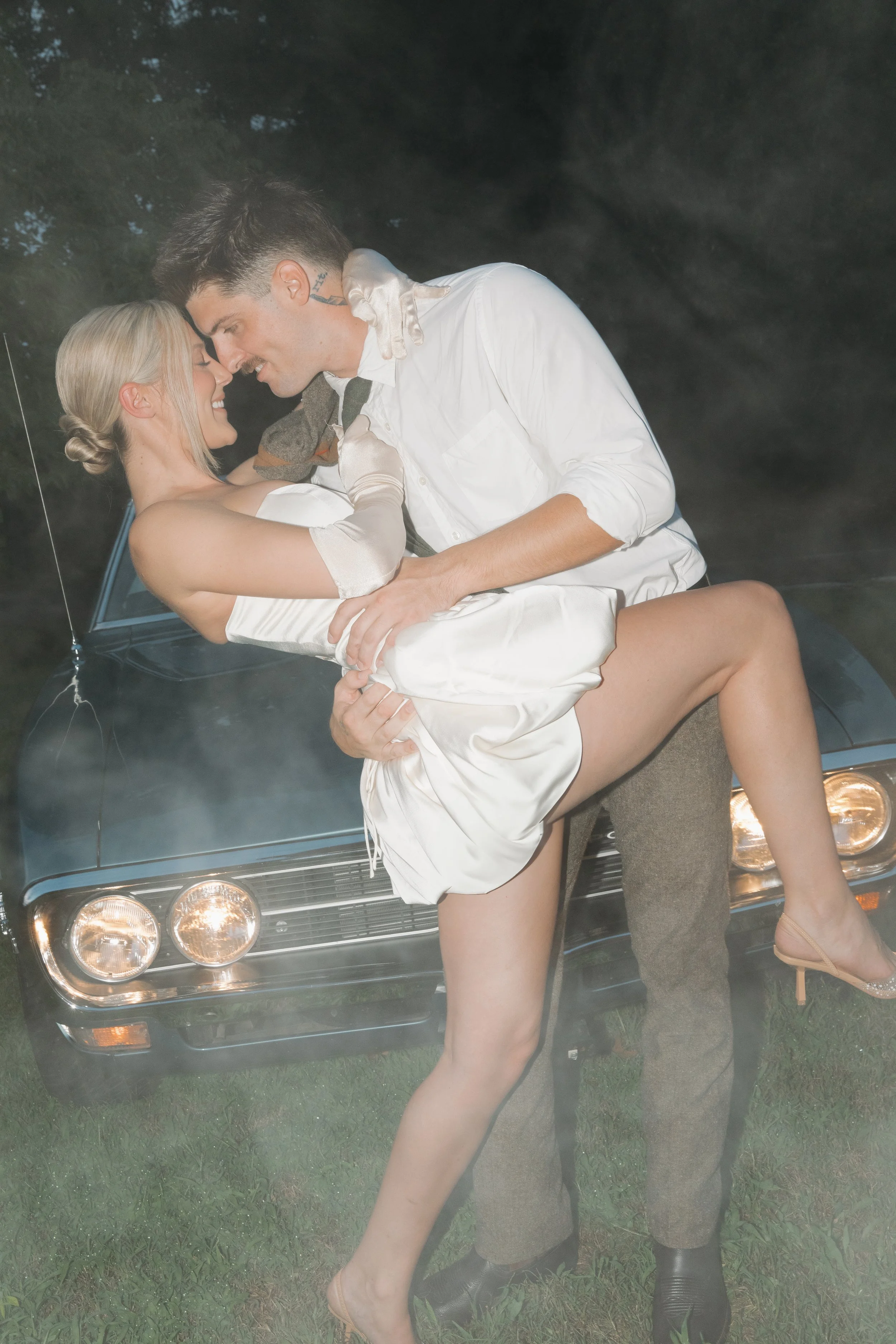 A man and woman are embracing and touching foreheads in front of a classic car with visible headlights, at night or dusk. The woman is wearing a satin dress and high heels, and the man is dressed in a white shirt and brown pants, with a scarf around his neck.