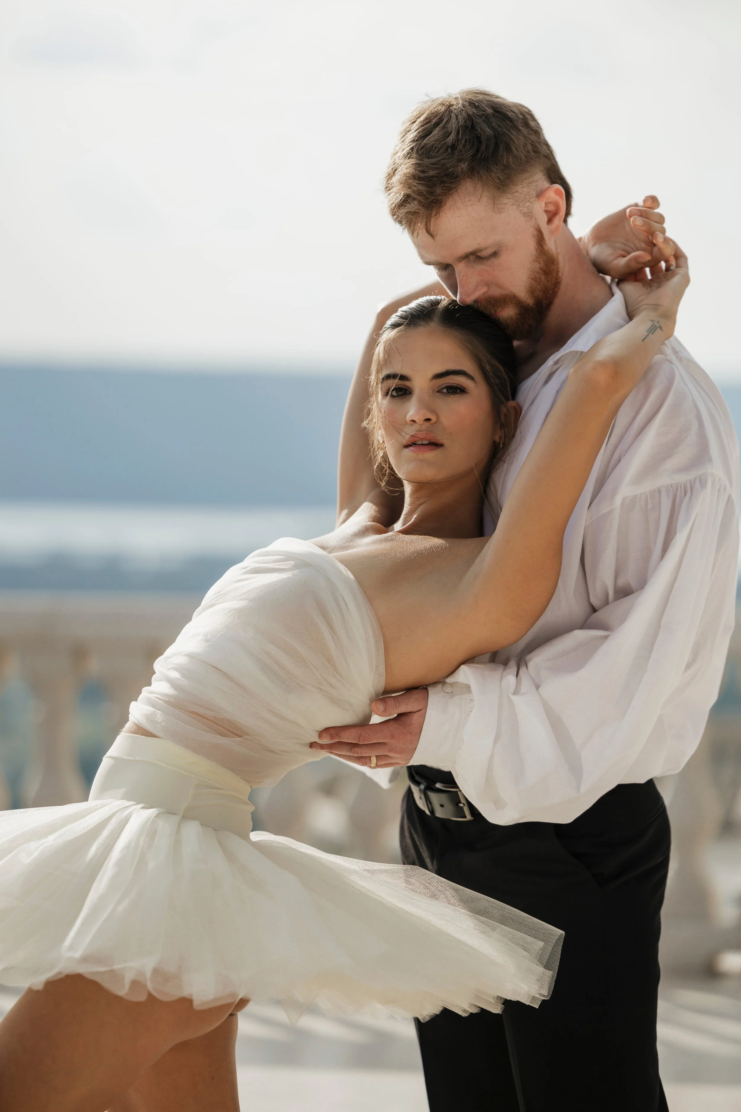 A man and woman embrace in a dance, with the woman wearing a white dress with a ruffled skirt and the man in a white shirt and black pants.