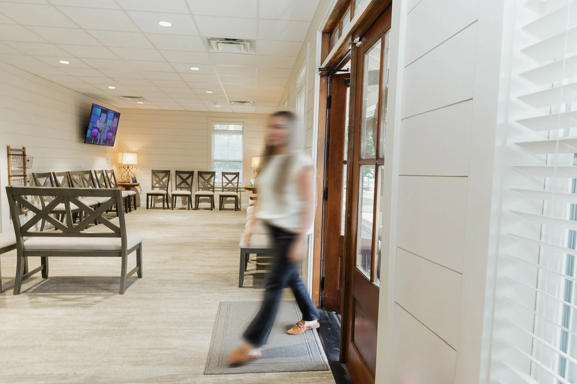 Blurred woman entering a room through a wooden door with glass panels, beige walls, and a television mounted on the wall in a waiting room with chairs and lamps.
