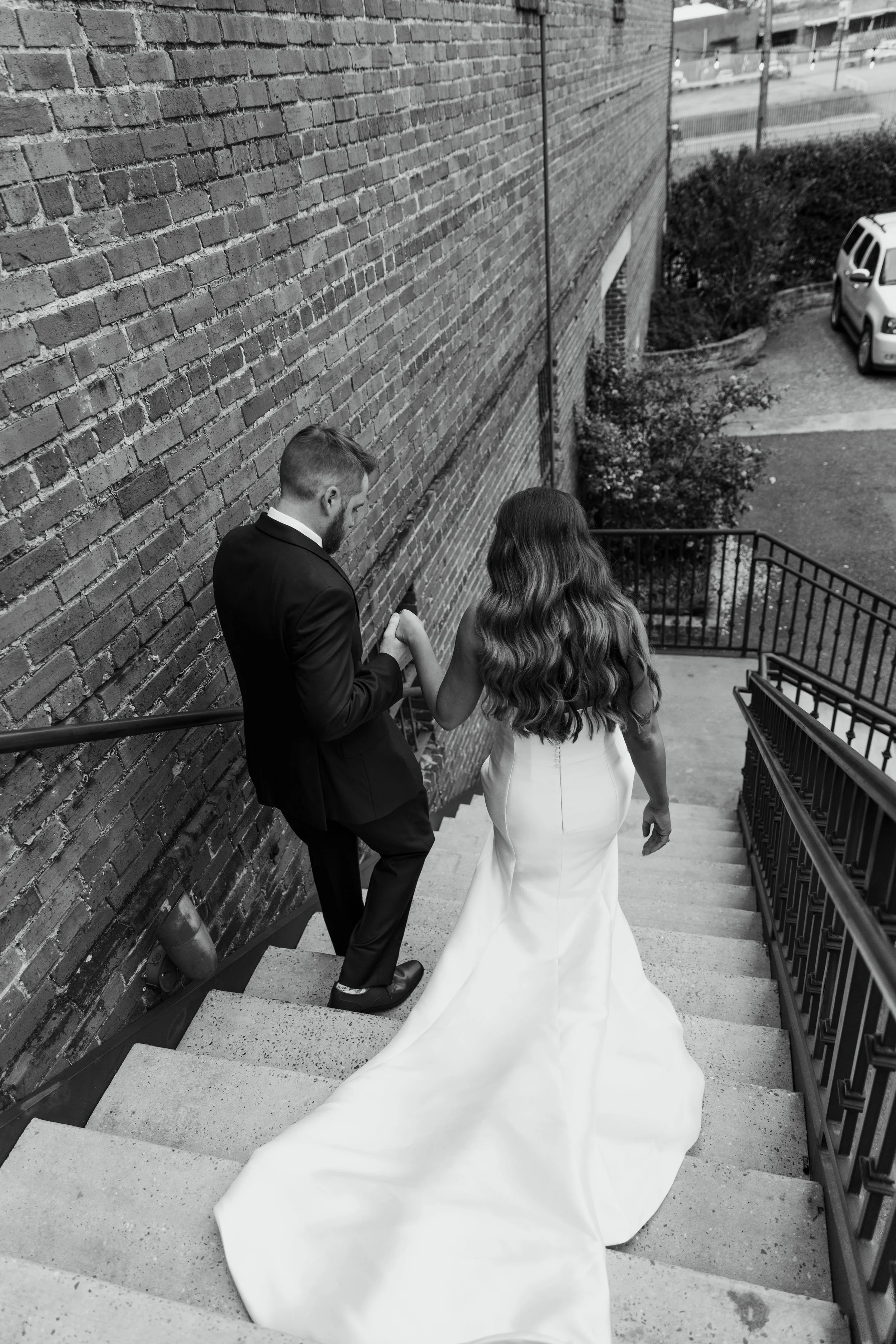 A bride and groom holding hands ascending outdoor concrete stairs beside a brick wall, with the bride in a long white wedding dress and the groom in a dark suit.