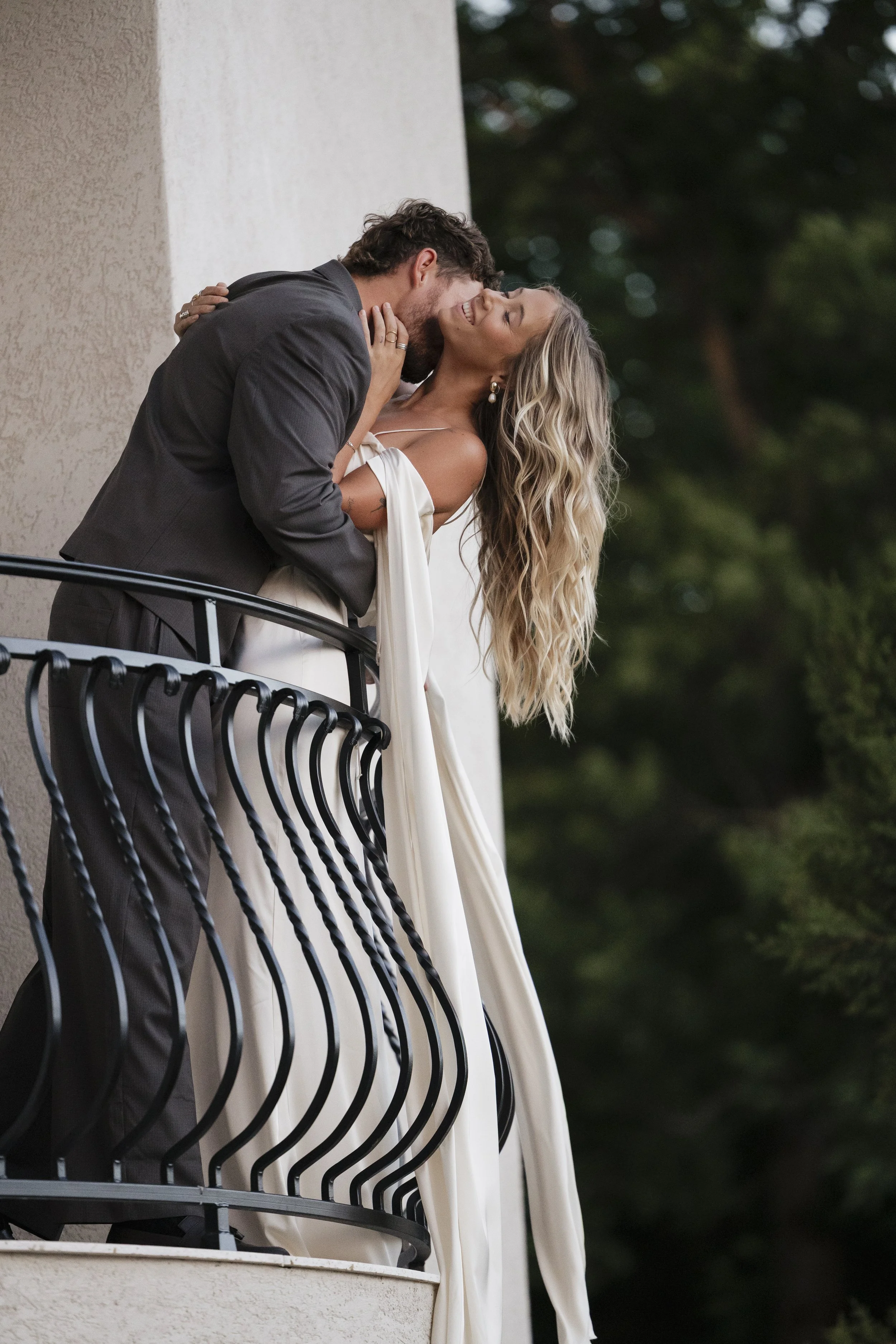A couple embraces and shares a kiss on a balcony with a black wrought iron railing, with greenery in the background.