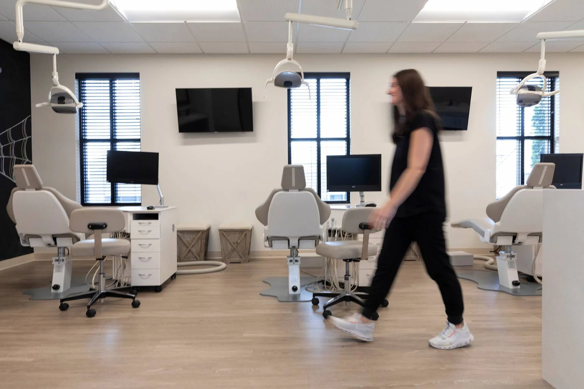 A woman walking in a dental clinic with reclining dental chairs, computer monitors, and medical equipment, in a well-lit room with large windows and wooden flooring.