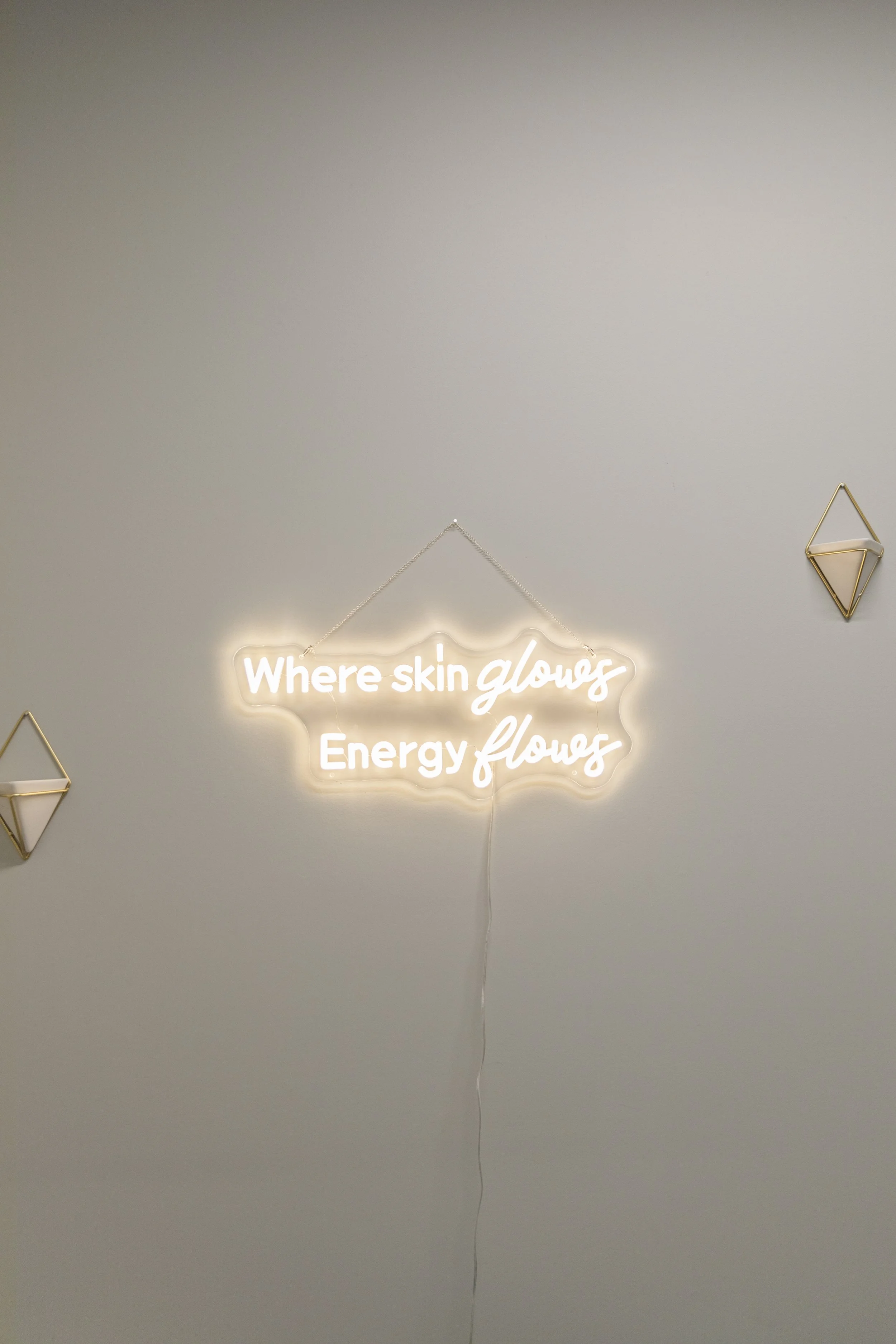 Neon sign on wall reading 'Where skin glows, Energy flows' with decorative gold geometric wall accents nearby.