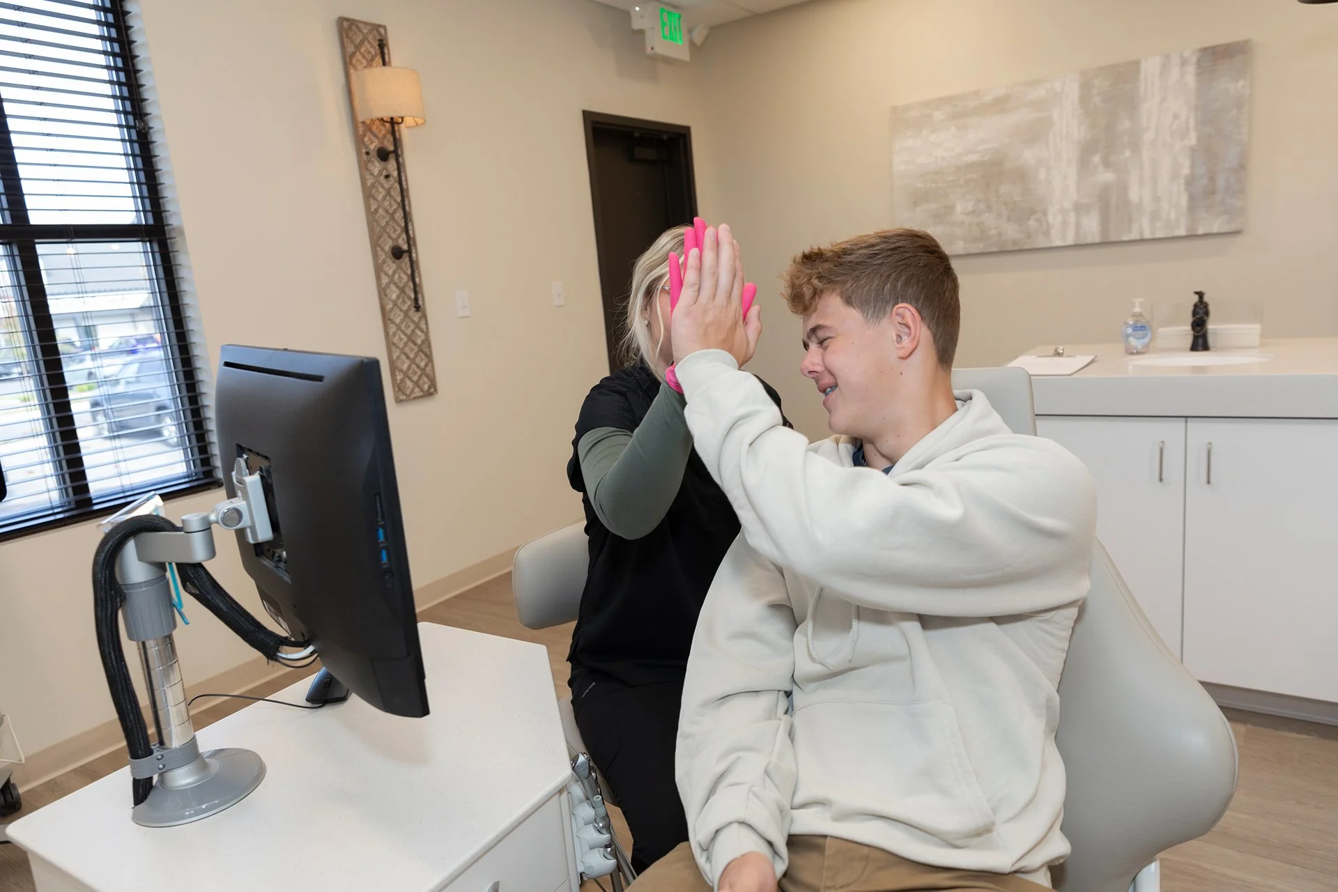 A young man sitting in a chair at a healthcare facility, smiling and giving a high-five to a nurse or medical professional who is standing next to him. There is medical equipment and a monitor on the desk in front of him, and the room has a large window with blinds, a wall light, and hanging decor.
