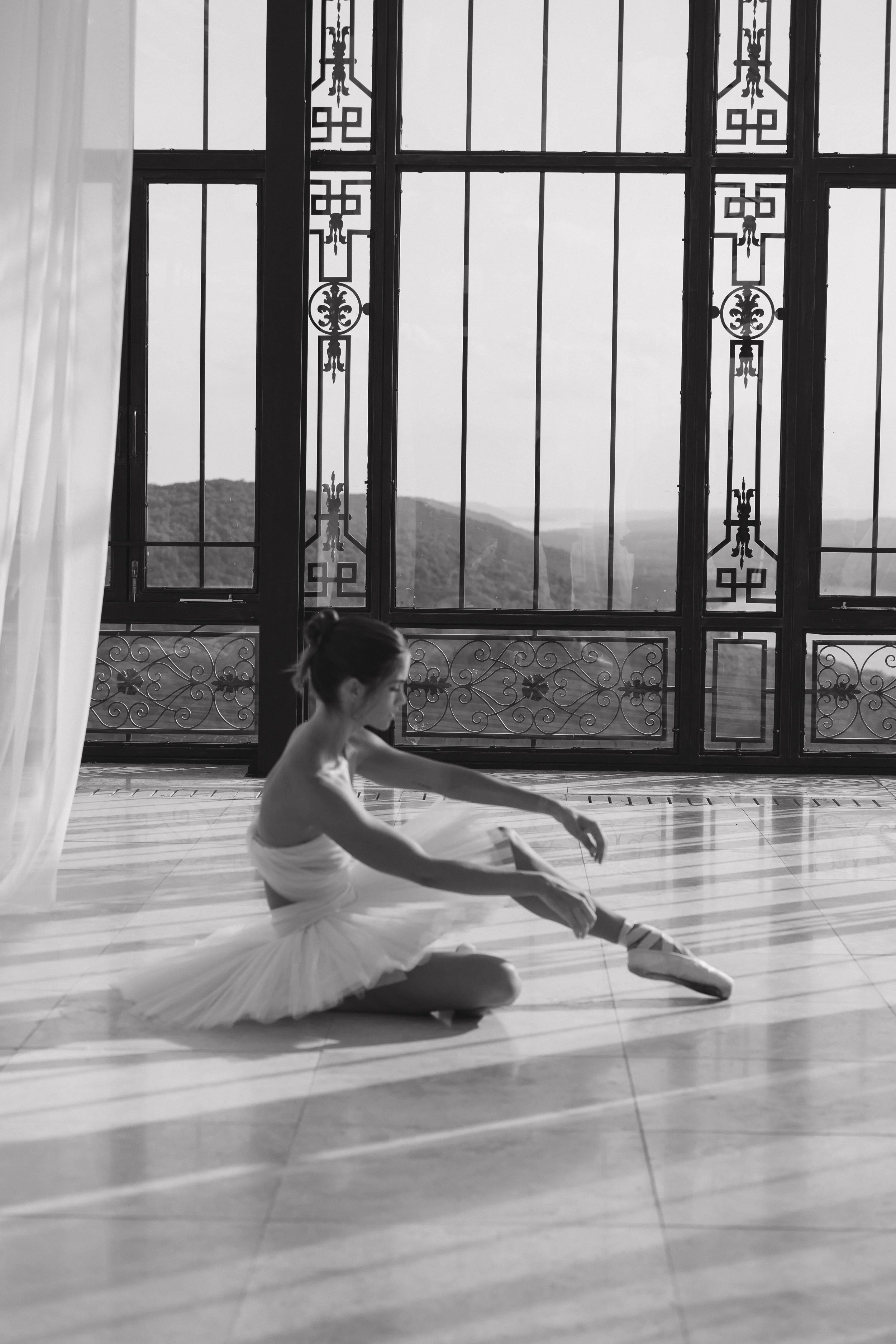 Black and white photo of bride seated on tile floor tying her pointe shoe ribbons in front of wrought iron windows and white drapery at Stone Haven Weddings Alabama, classic ballerina bridal editorial by Erin Daniel, middle Tennessee wedding photogra