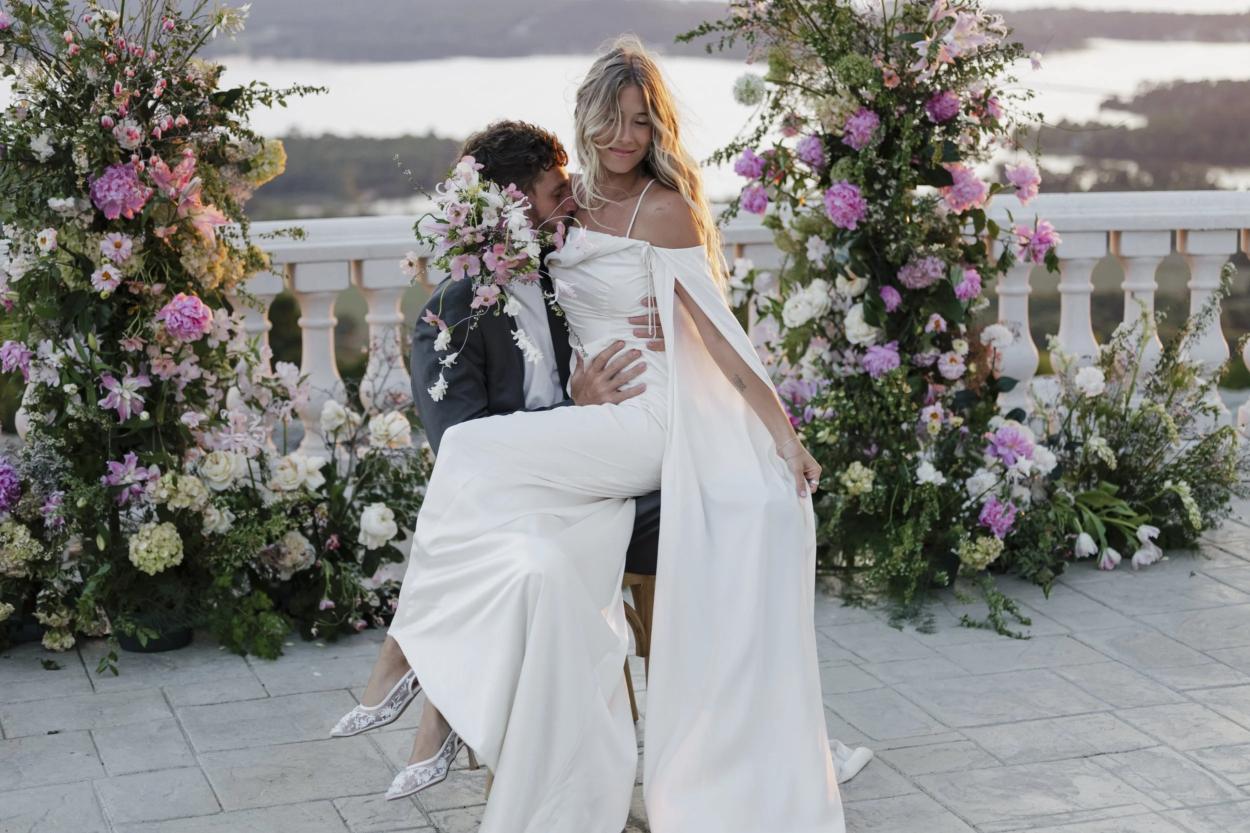 A couple at their wedding, with the groom sitting and the bride standing, surrounded by an arch of pink and white flowers, on a patio overlooking a body of water and distant hills.