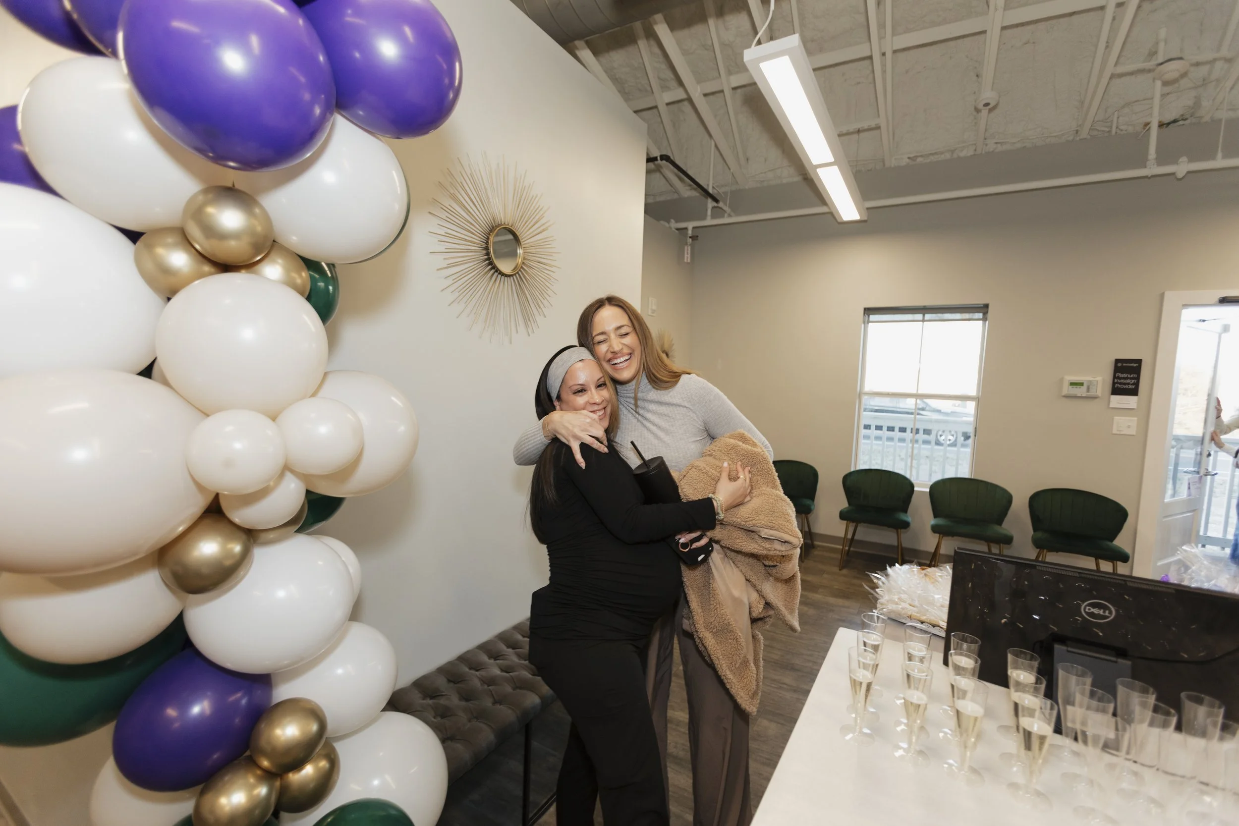 Two women hugging and smiling at a celebration with balloons and champagne glasses indoors.