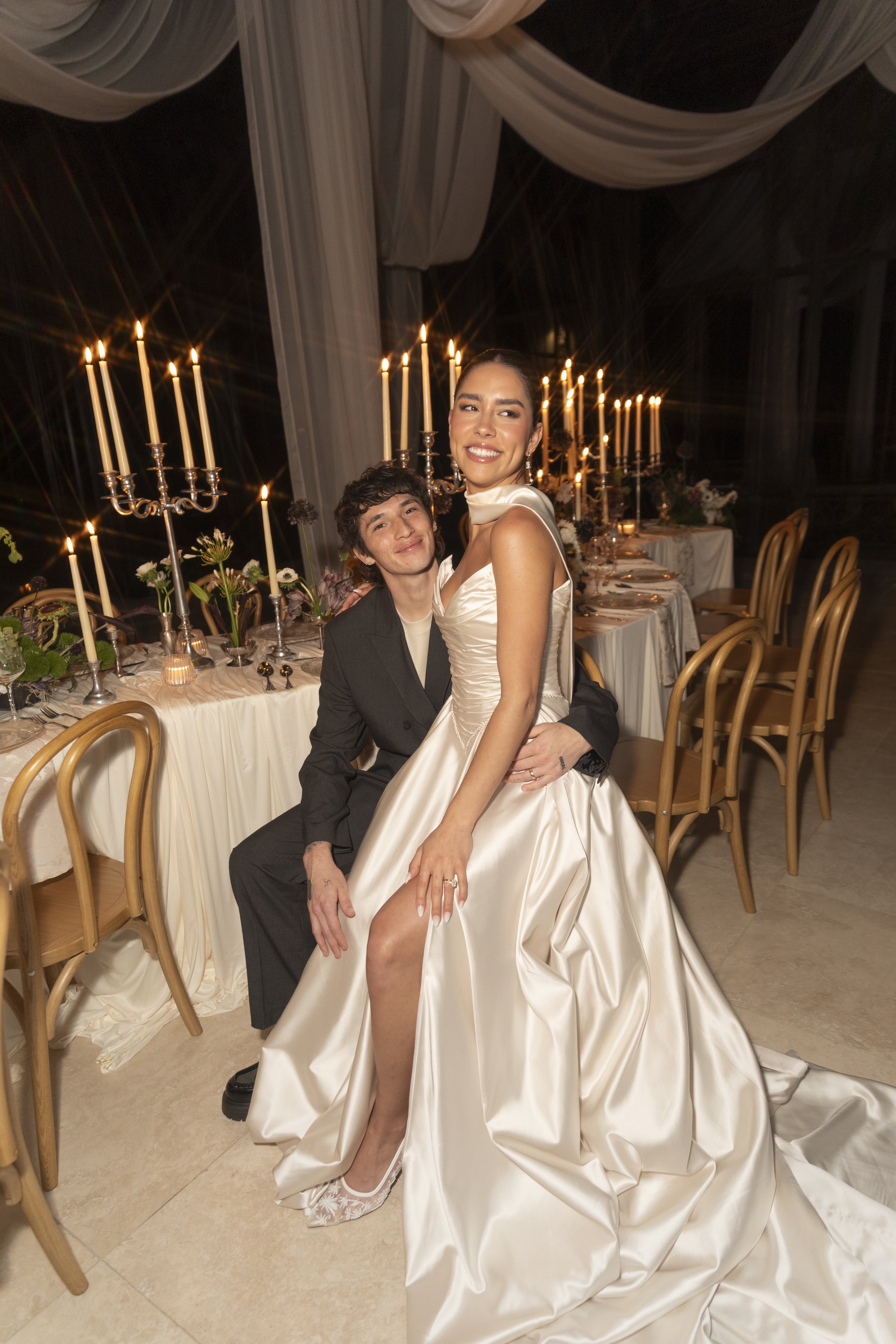 A woman in a satin wedding dress sitting on a man's lap at a formal dinner event, with a long table set with candles and flowers in the background.