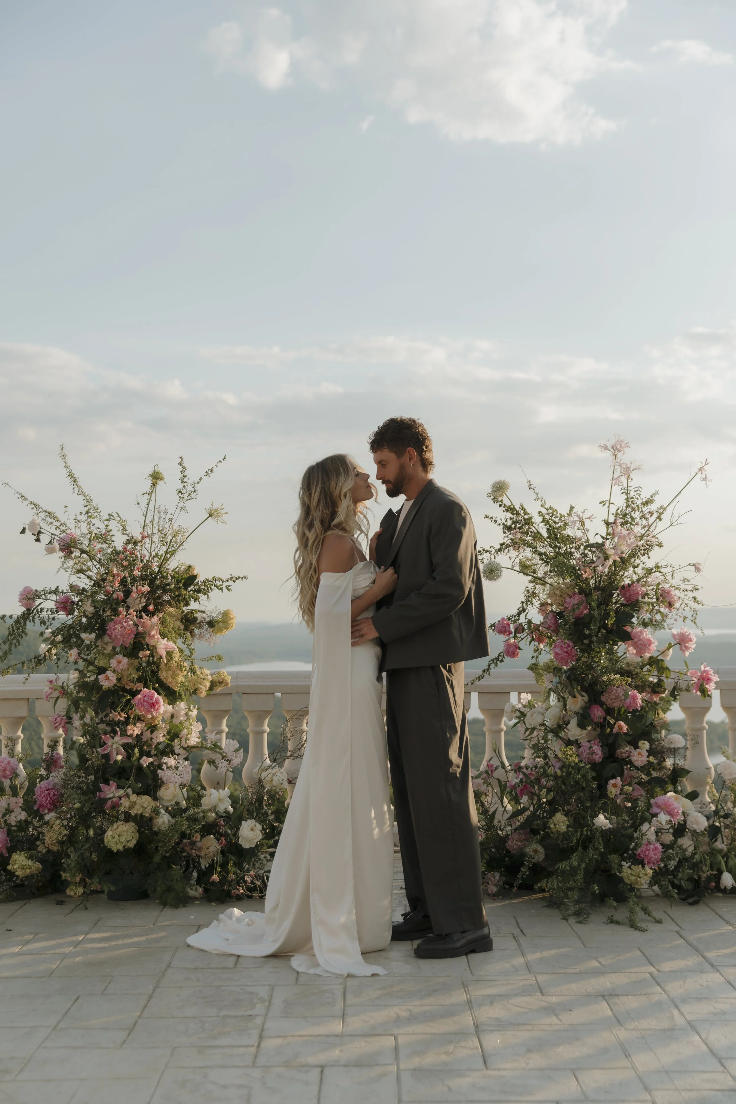 A bride and groom standing close together on a balcony with a scenic view of the sky and distant landscape, surrounded by two large arrangements of pink and white flowers.