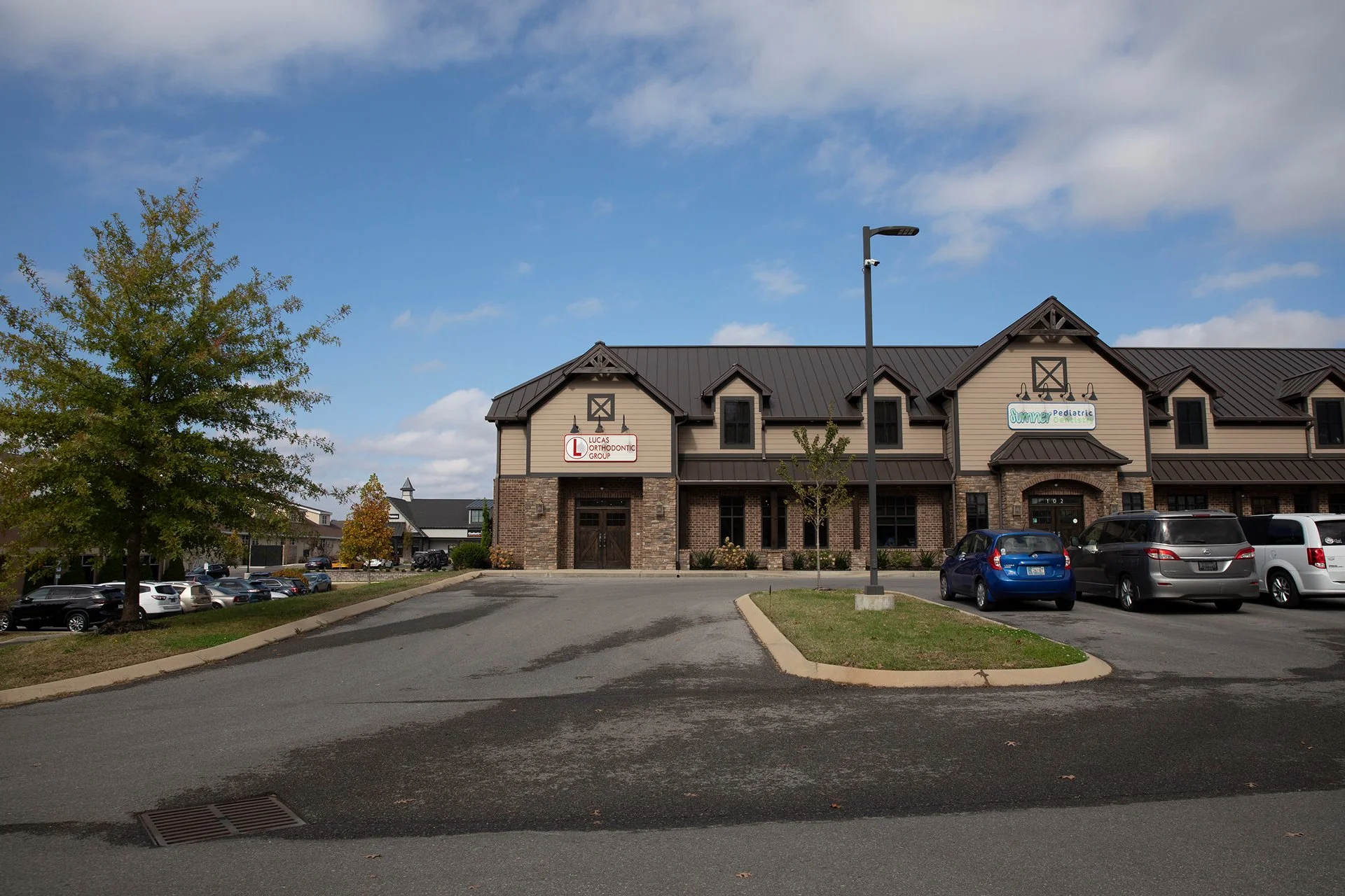 A commercial building with a brick and beige siding facade, said to house Lucas Orthodontic Group and Summer Pediatric Dentistry. There is a parking lot in front with several cars, a tree to the left, and a streetlight in the middle. The sky is partly cloudy.