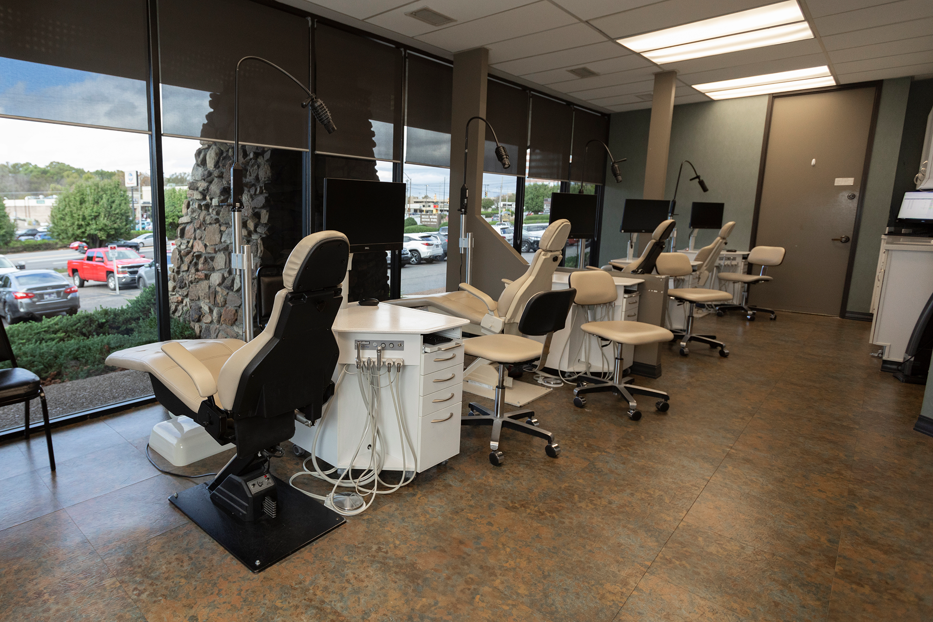Inside a dental clinic with dental chairs, monitors, and dental tools, large windows with view of parking lot and trees outside.