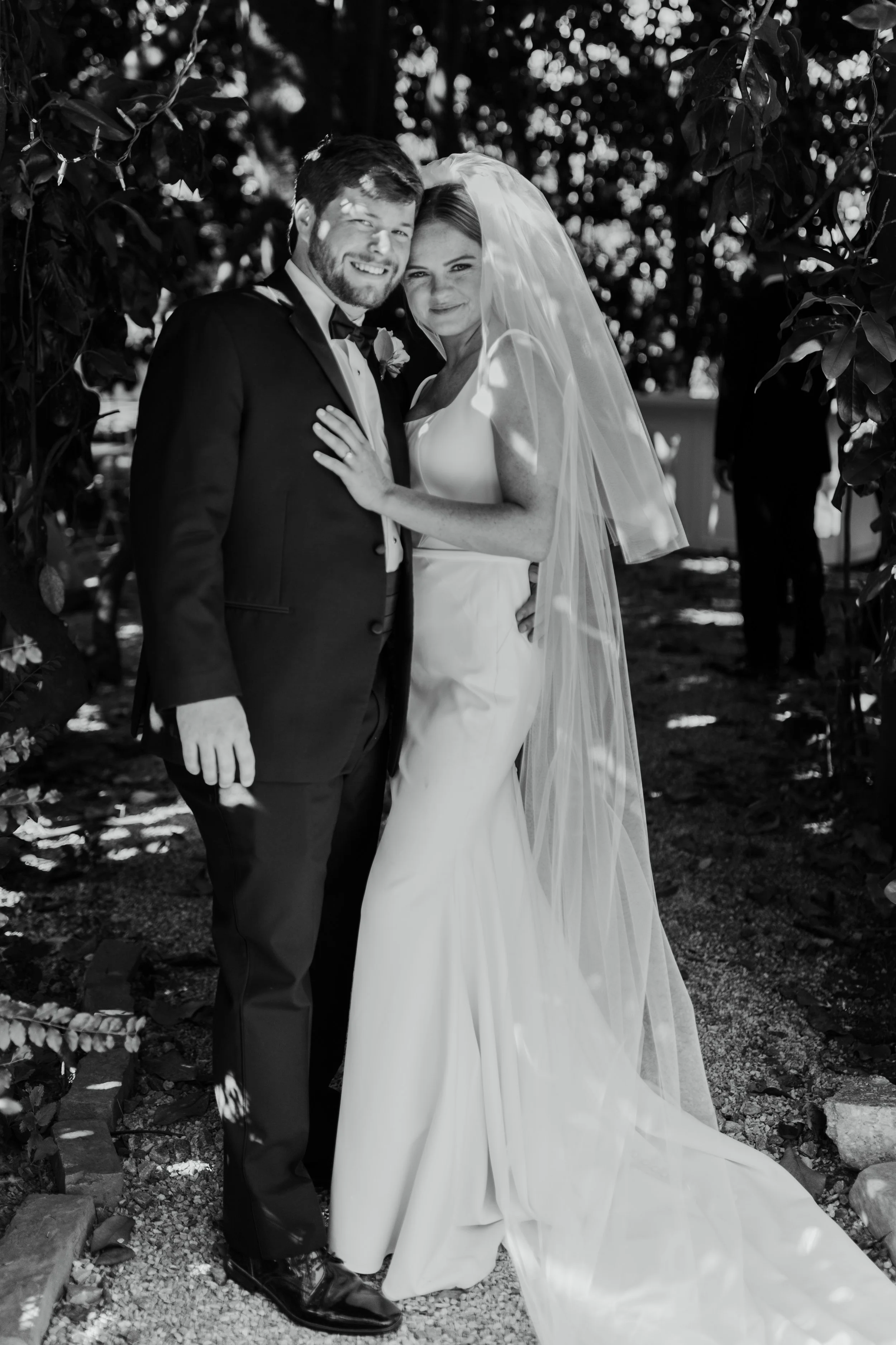 A bride and groom are smiling and embracing outdoors, dressed in wedding attire, with trees in the background.