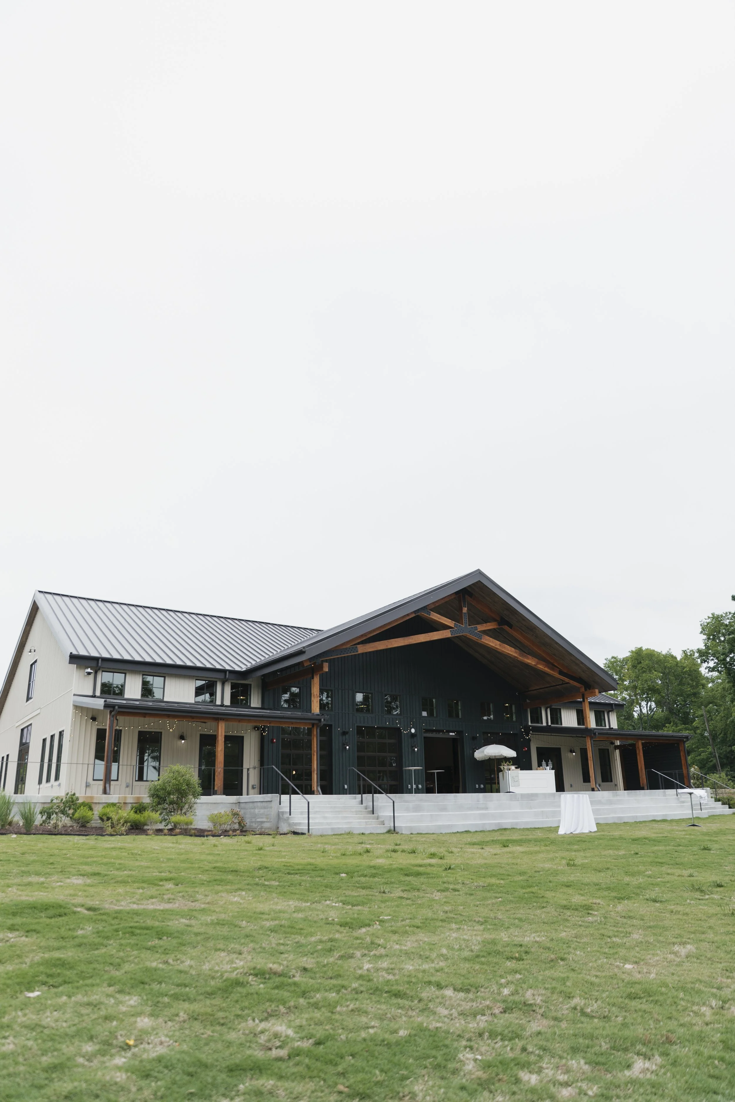 A large modern house with black and white exterior siding, a metal roof, and a spacious front porch area with steps and string lights, surrounded by a green lawn and some trees, under an overcast sky.