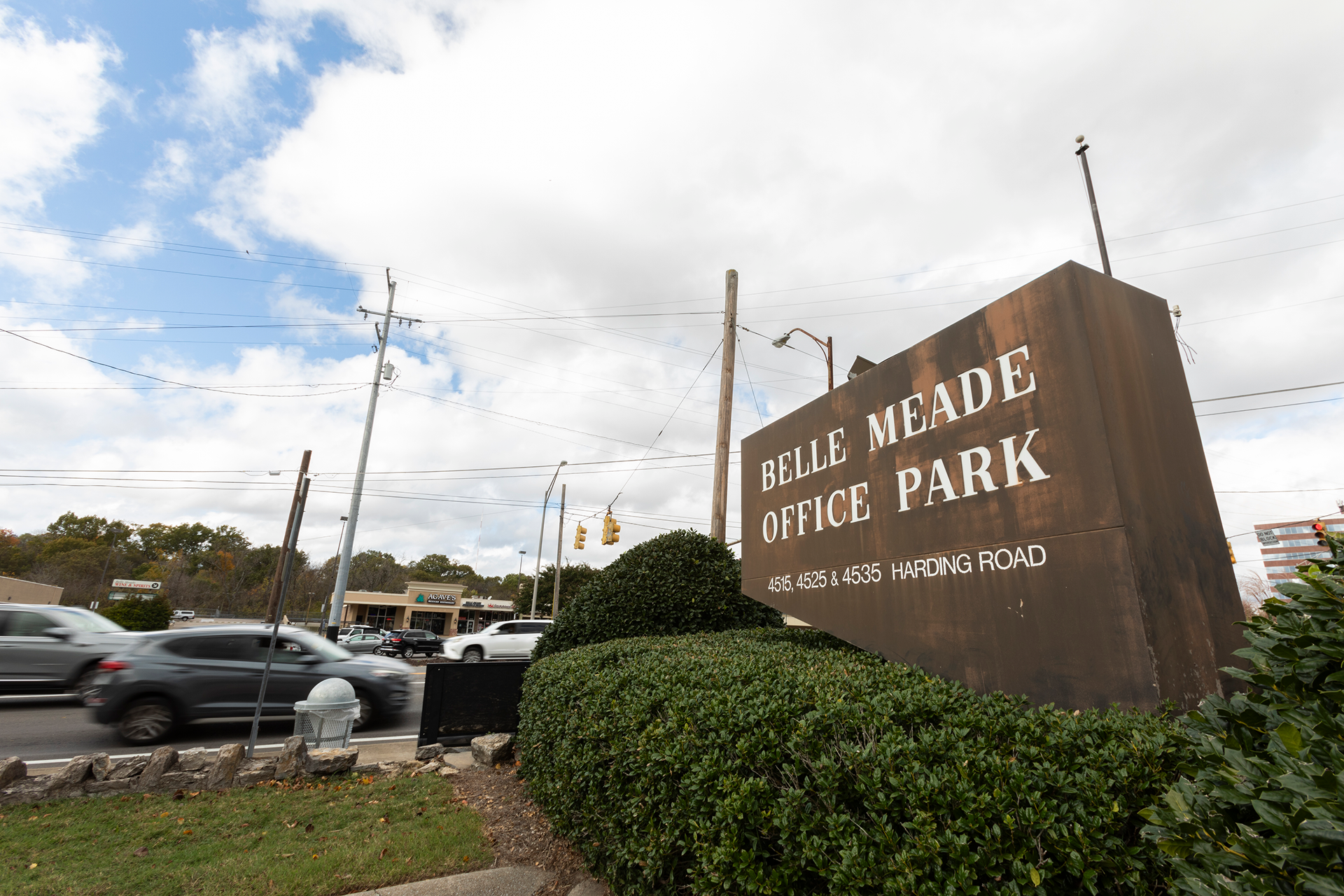 A large rust-colored sign for Belle Meade Office Park with white lettering, situated among green bushes on a street corner. In the background, there are cars driving past and power lines against a partly cloudy sky.