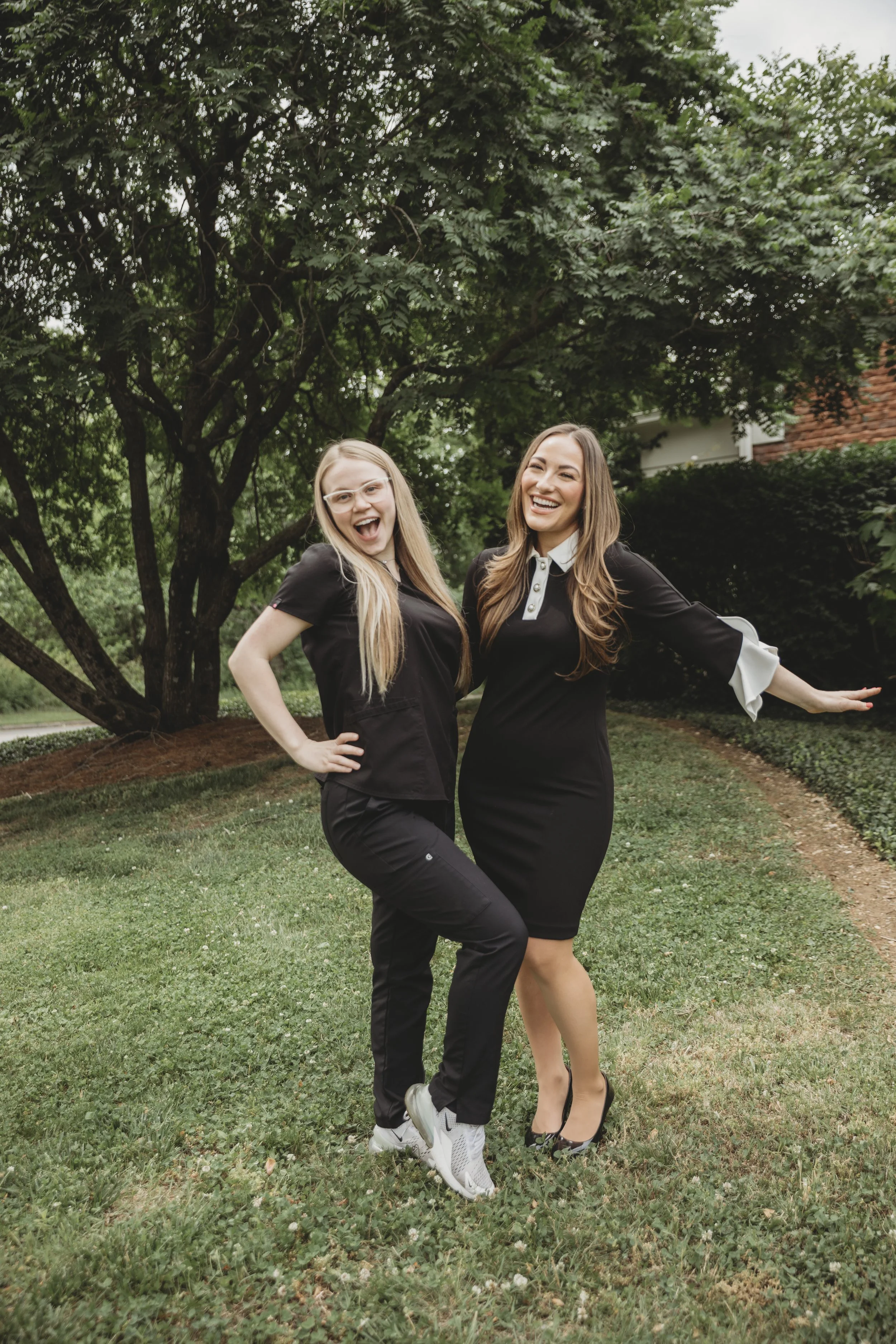 Two women in black outfits, smiling and posing together outdoors near a large tree on a grassy area with bushes and a house in the background.