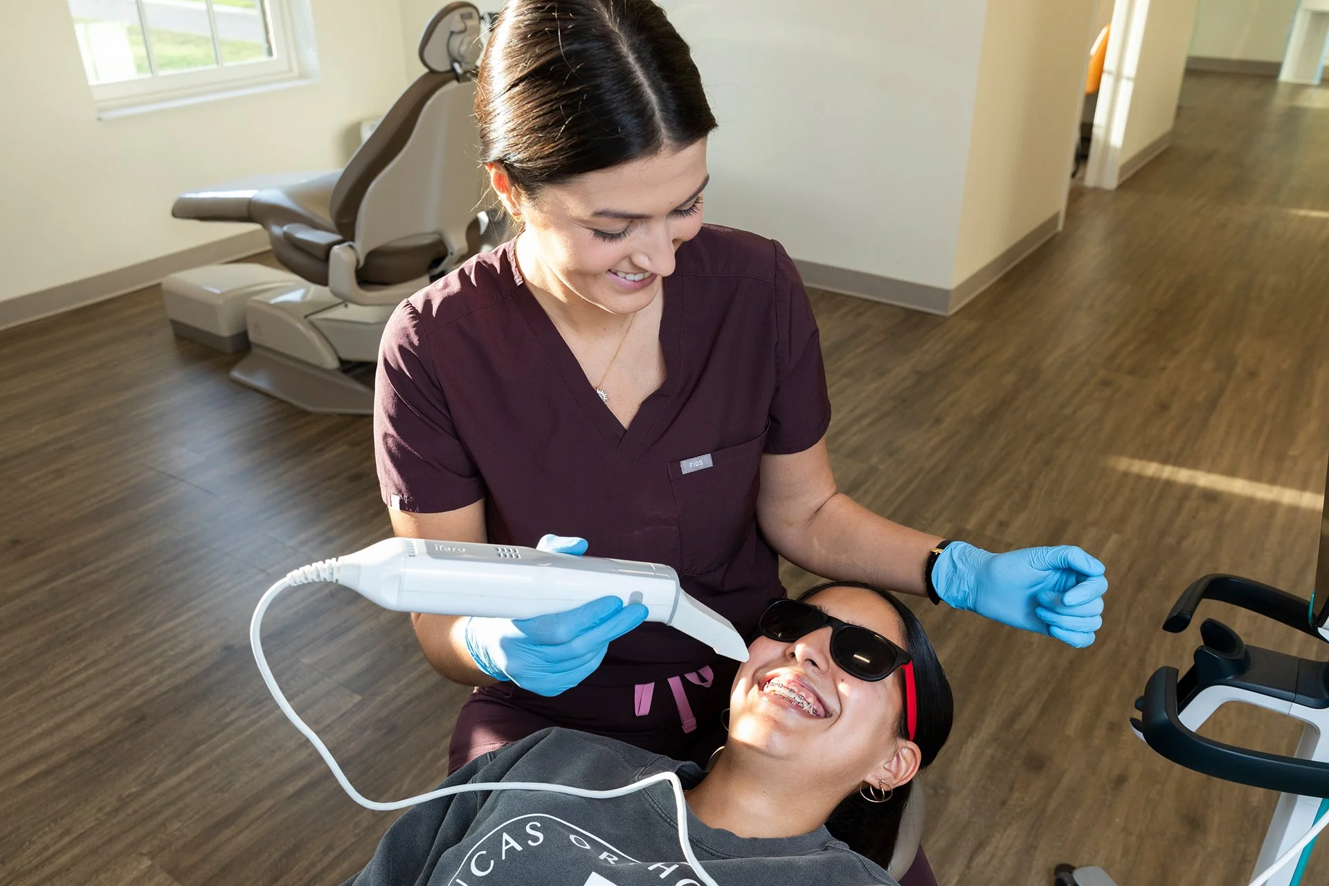 A dentist in maroon scrubs and blue gloves examines a patient's teeth with a dental instrument in a well-lit dental office. The patient, wearing sunglasses, is smiling. In the background, there is a dental chair near a window.